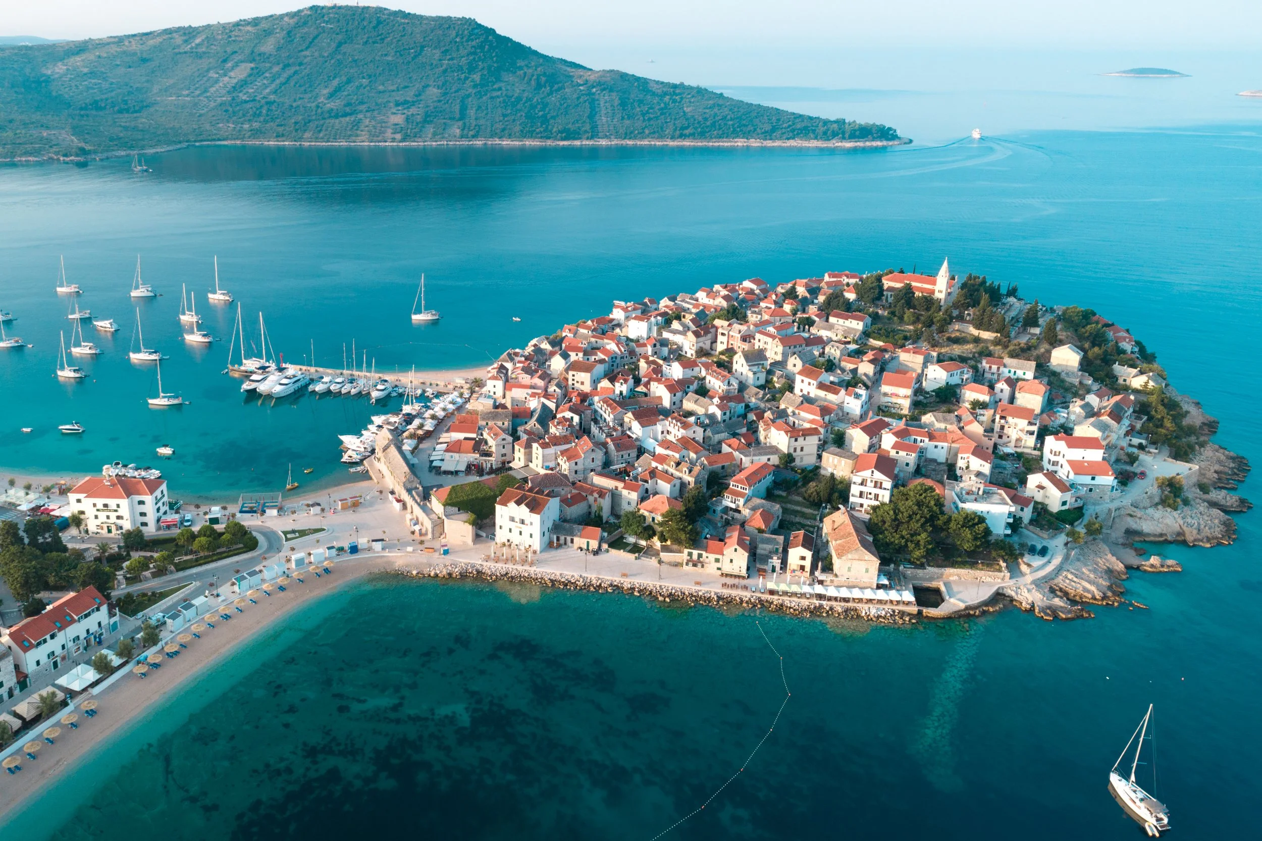 An aerial view of a small coastal town on a peninsula with red-roofed buildings, a marina with sailboats, and surrounding turquoise waters and hilly landscape.
