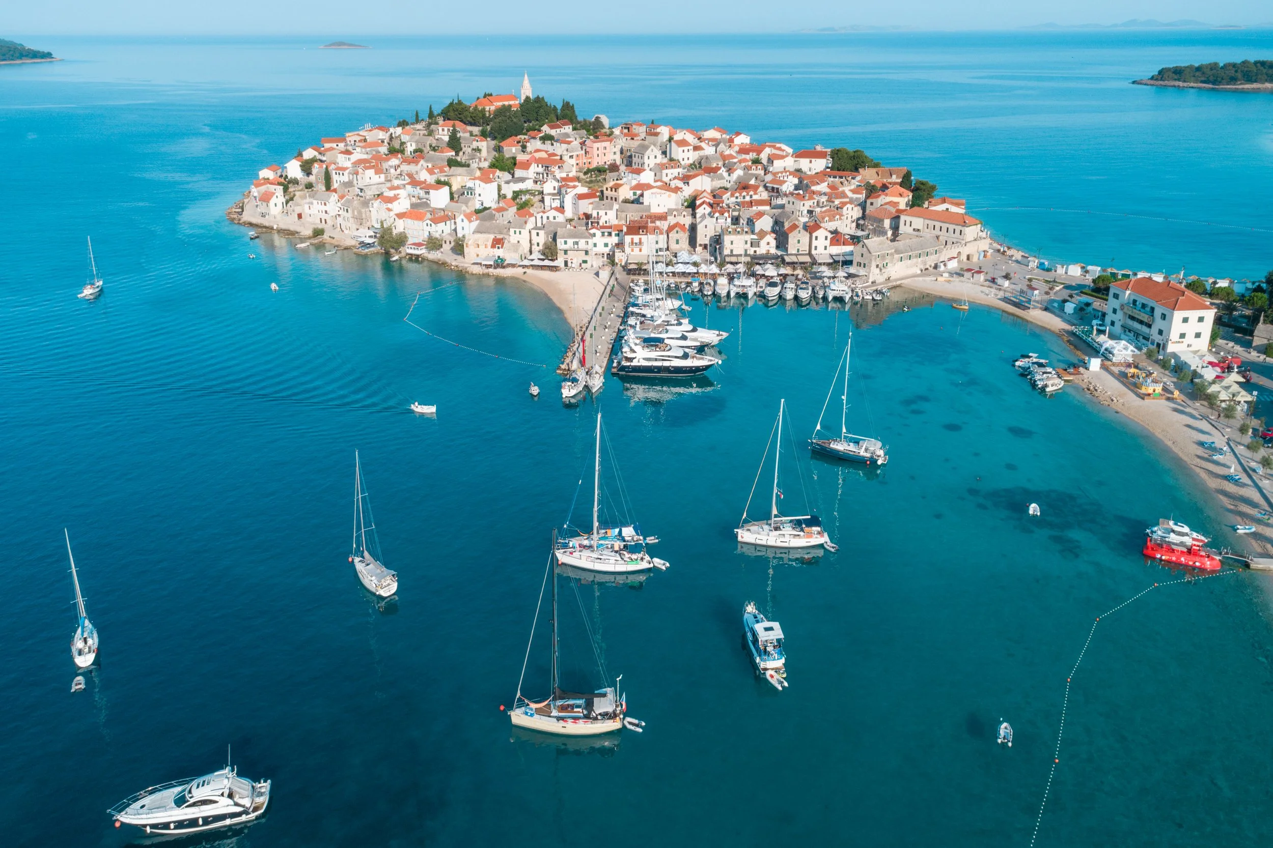Aerial view of a coastal town on an island with a harbor filled with sailboats, surrounded by blue water and open sea in the background.