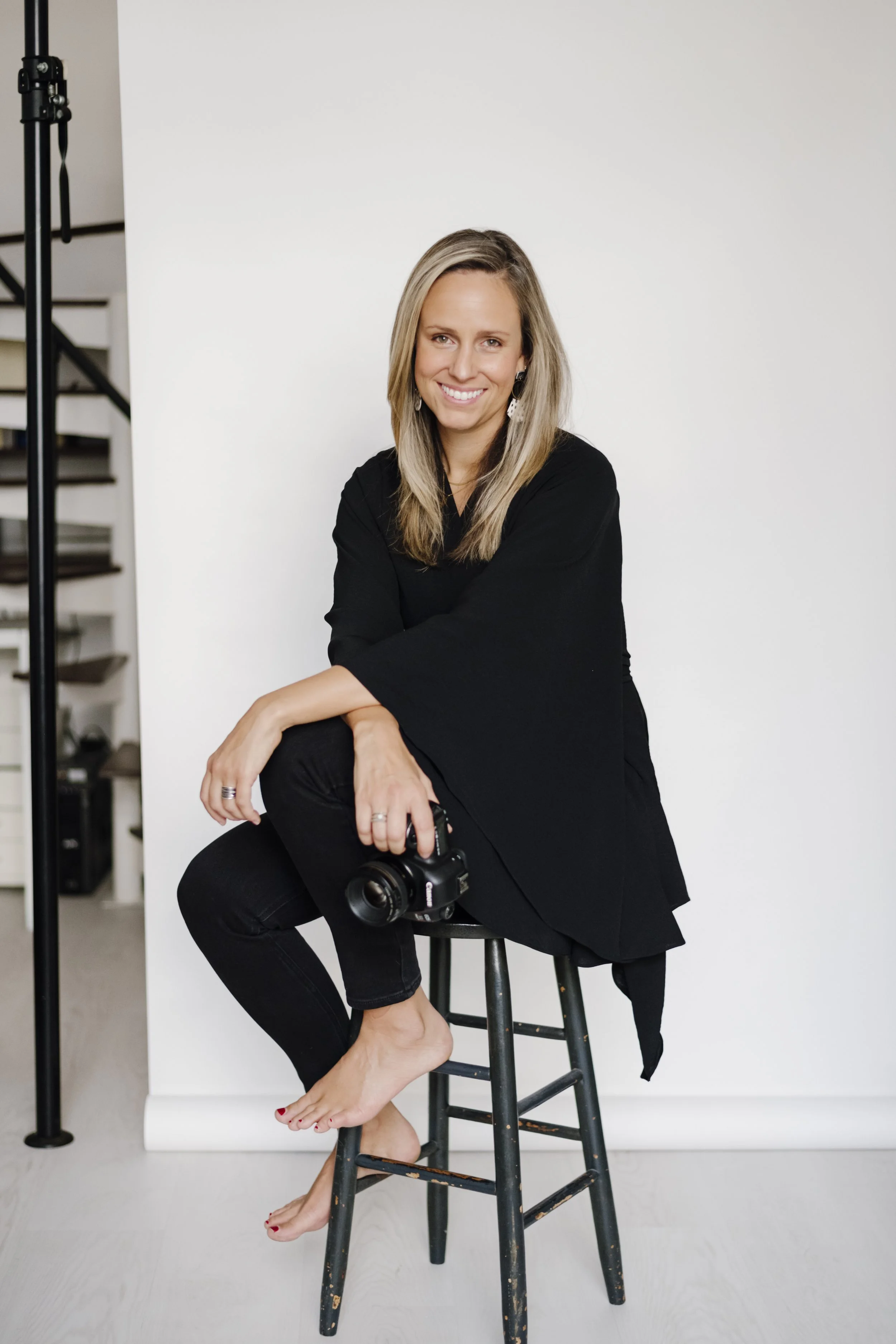 Self portrait. JJ Ignotz sitting on a stool holding a camera in a studio setting.