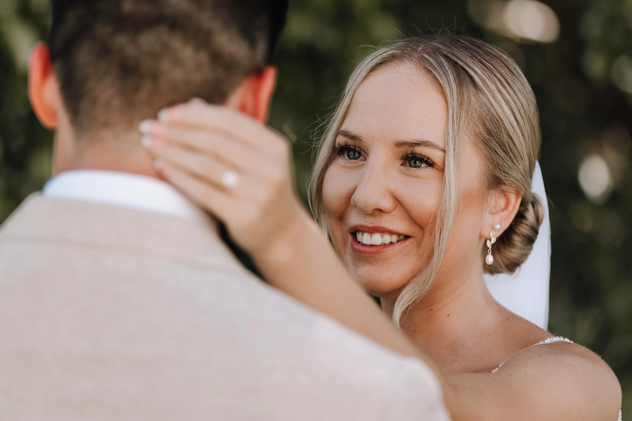 A bride and groom embrace outdoors during their wedding, with the bride's face visible as she smiles at the groom.