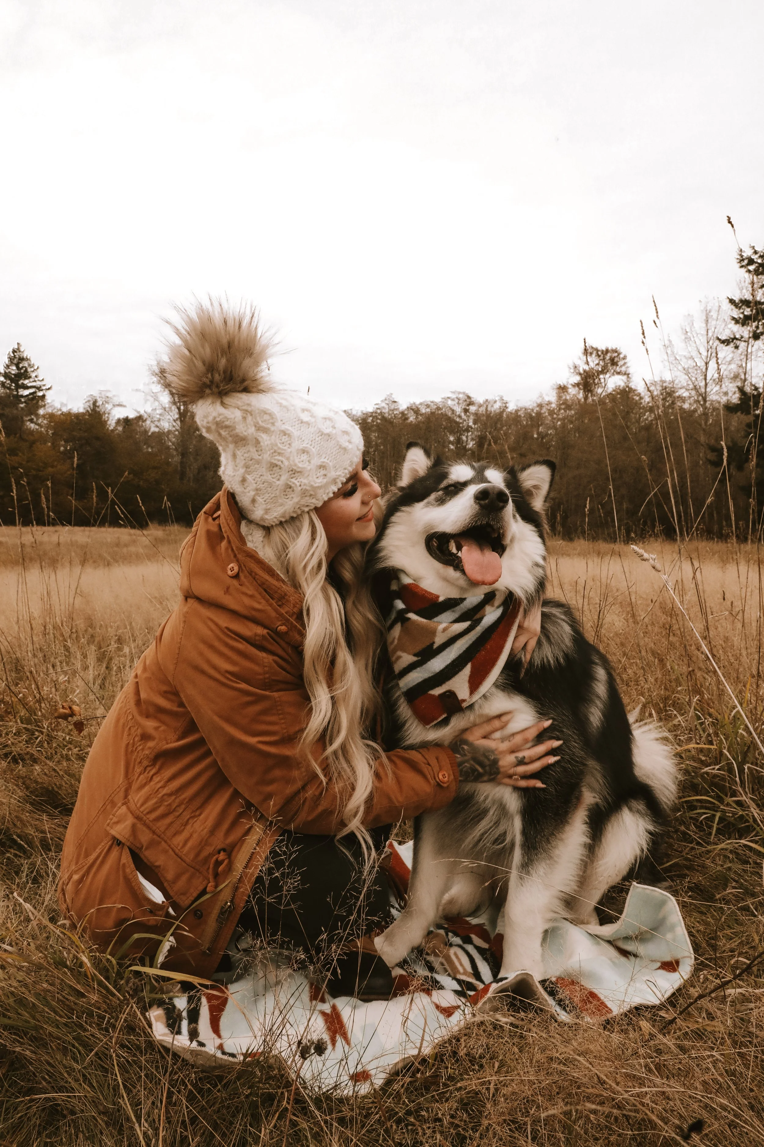 A woman hugging a happy Siberian Husky dog in a field, wearing a scarf, jacket, and knit hat with a pom-pom during autumn.