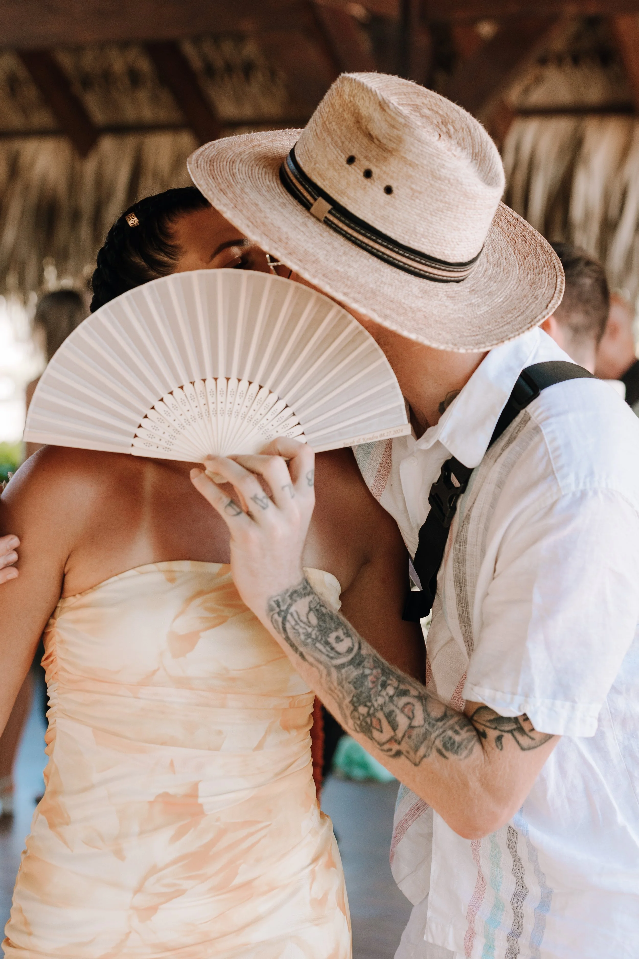 A couple sharing an intimate moment, with the woman holding a fan covering part of her face and the man wearing a straw hat, at a tropical setting.