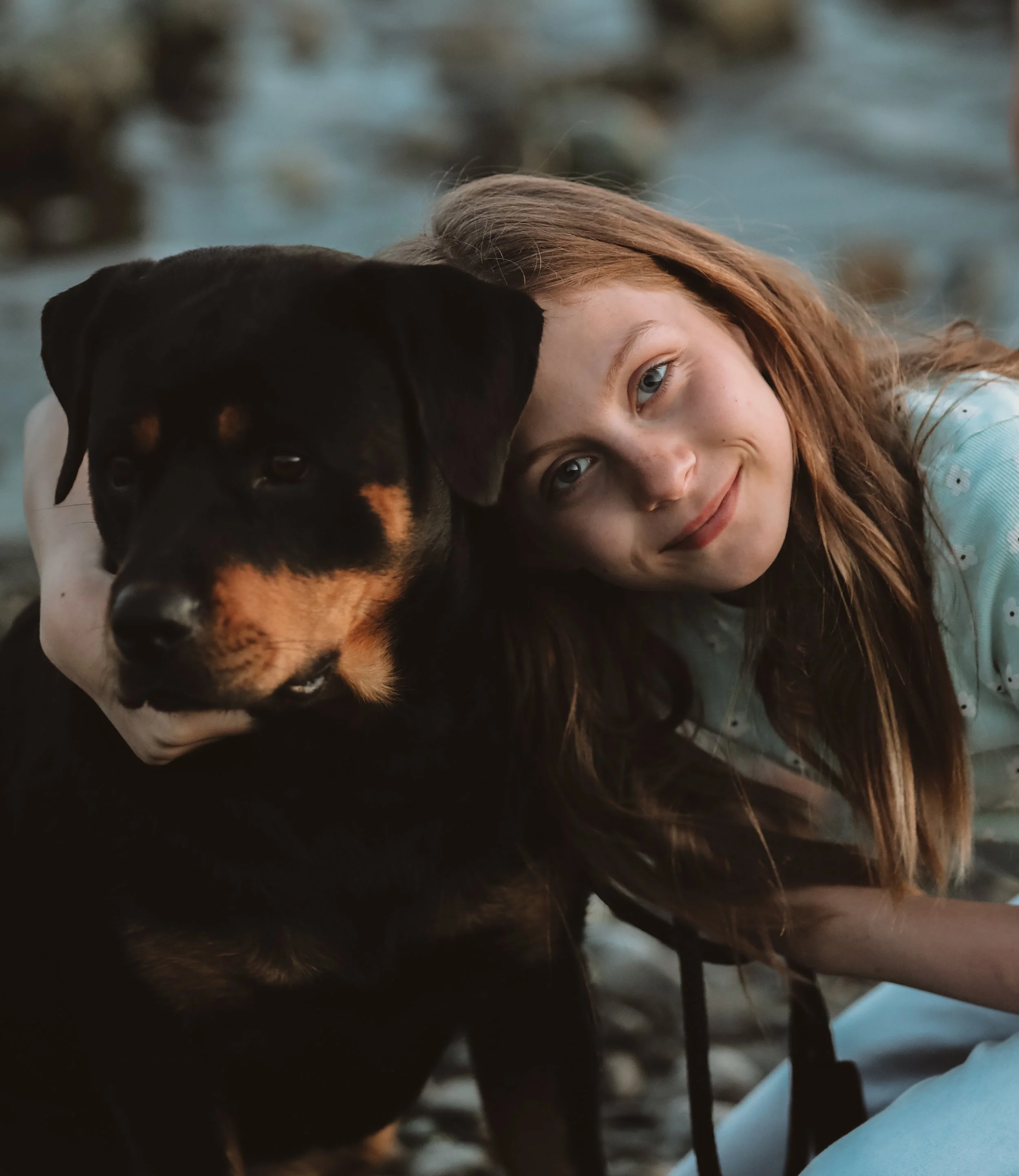 A girl with long brown hair and a light blue shirt resting her head on a black and tan dog, both lying outdoors near a body of water.