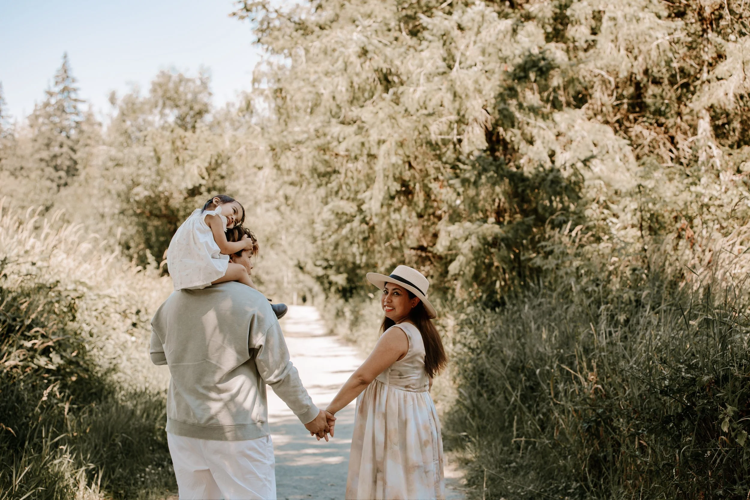 A family of three holding hands and walking on a nature trail, with a woman wearing a hat, a man in a hoodie carrying a girl on his shoulders, smiling amidst greenery and trees on a sunny day.