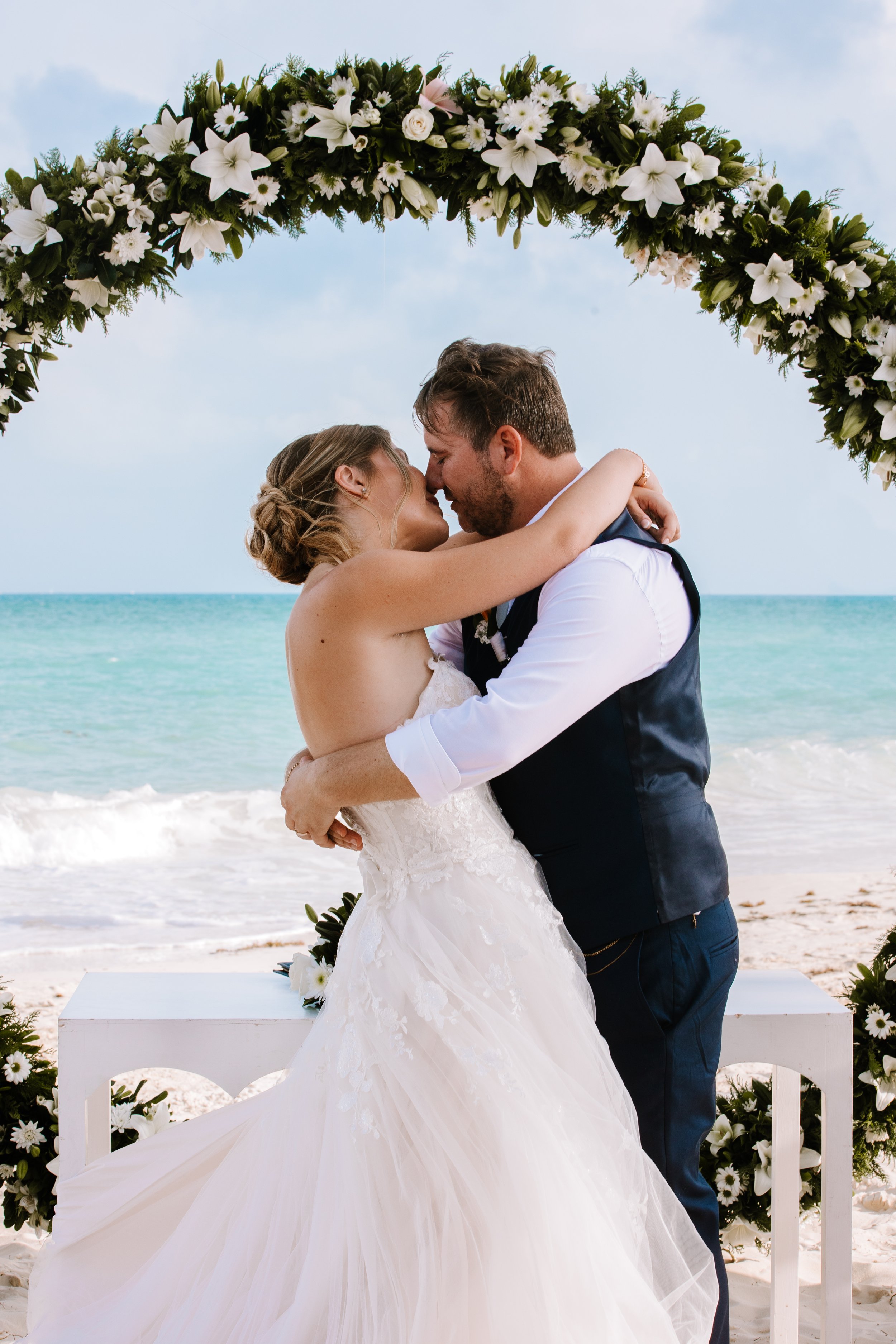A couple getting married on the beach, embracing under a floral arch with the ocean in the background.
