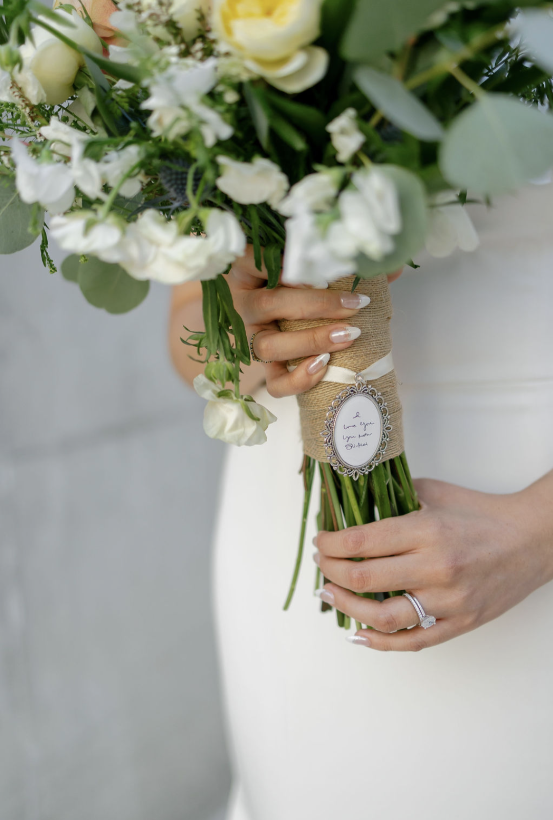A bride holding a bouquet of white and cream flowers with green leaves, wrapped in twine and decorated with a small silver frame and a note that says 'I love you, you make me smile.'