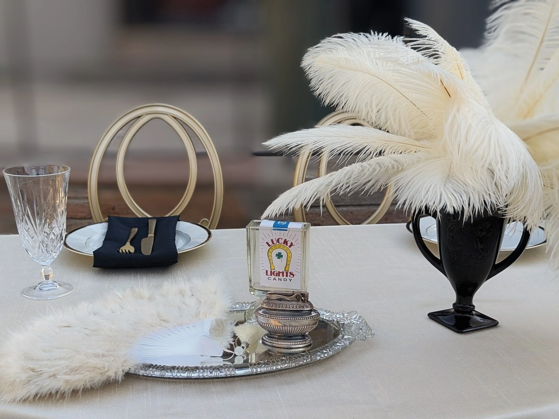 Elegant table setting with two clear glass goblets, a white plate with a black napkin and gold fork and knife, a silver tray with a vintage matchbook labeled 'Lucky Lights Candy,' and a black vase filled with white feathers. The background shows three cream-colored chairs with oval backs.