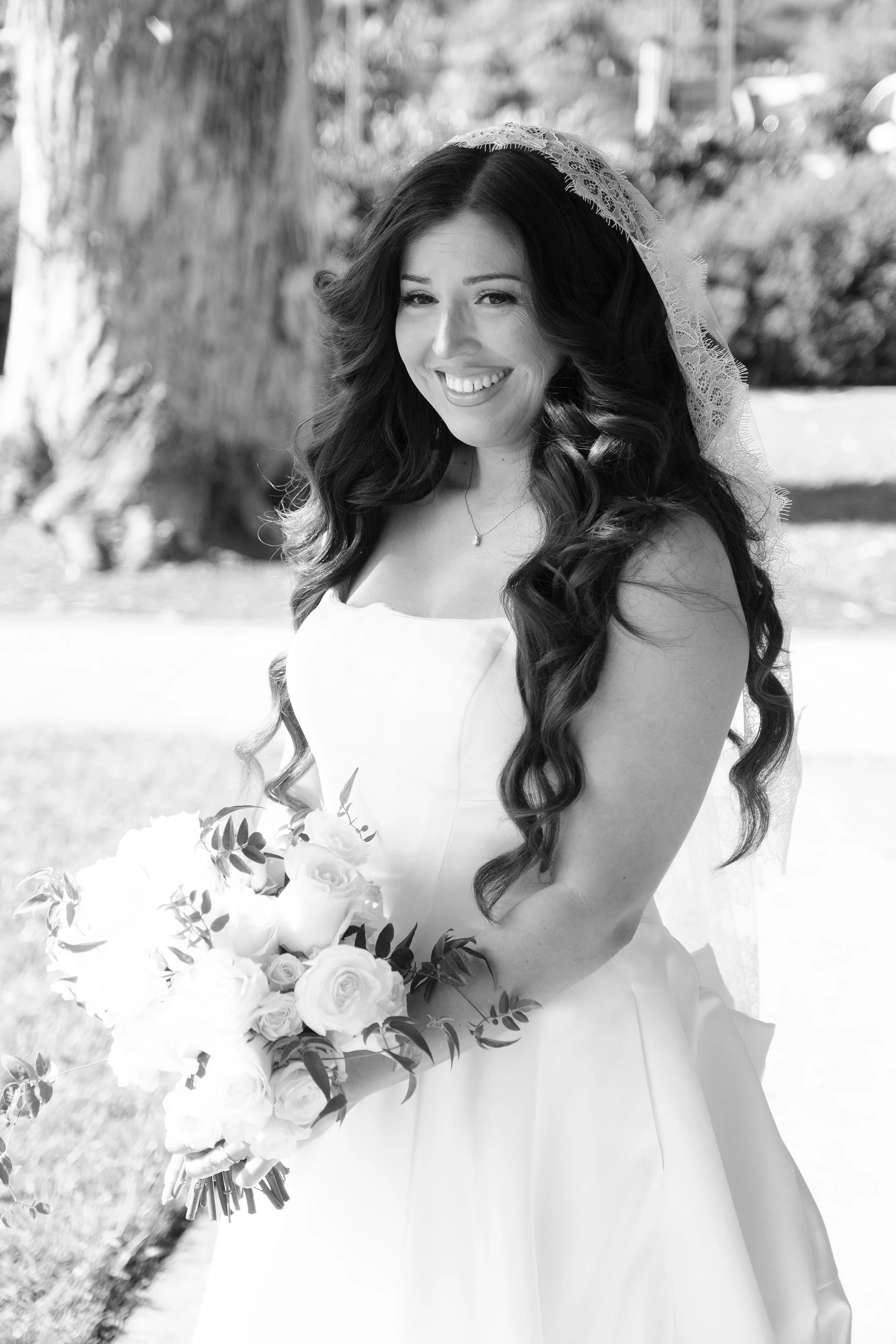 A woman in a wedding dress holding a bouquet of roses and greenery, with long wavy hair and a lace veil, smiling outdoors.