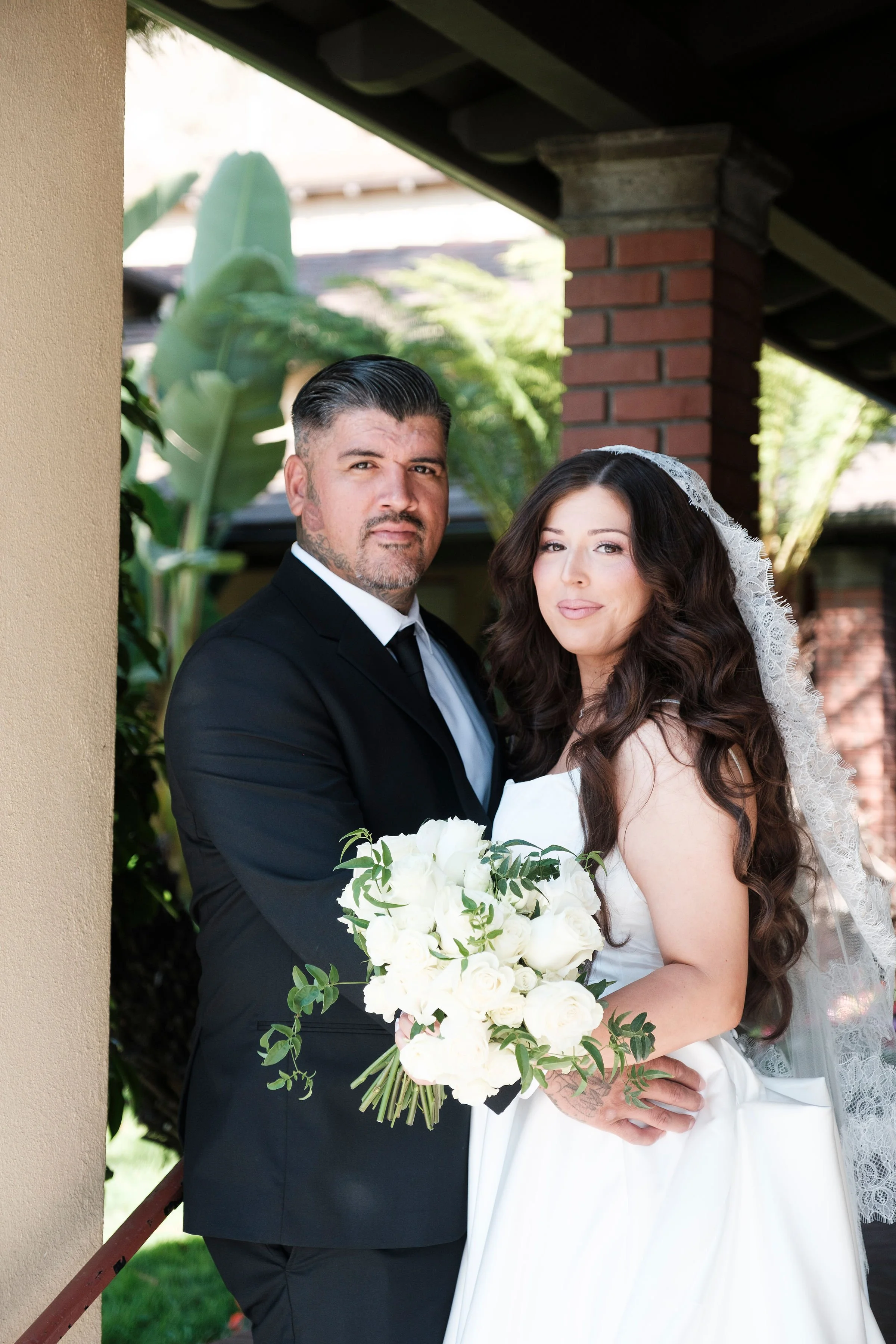 A groom and bride posing outdoors, with the groom in a black suit and tie, and the bride in a white wedding gown and veil, holding a bouquet of white roses and greenery.
