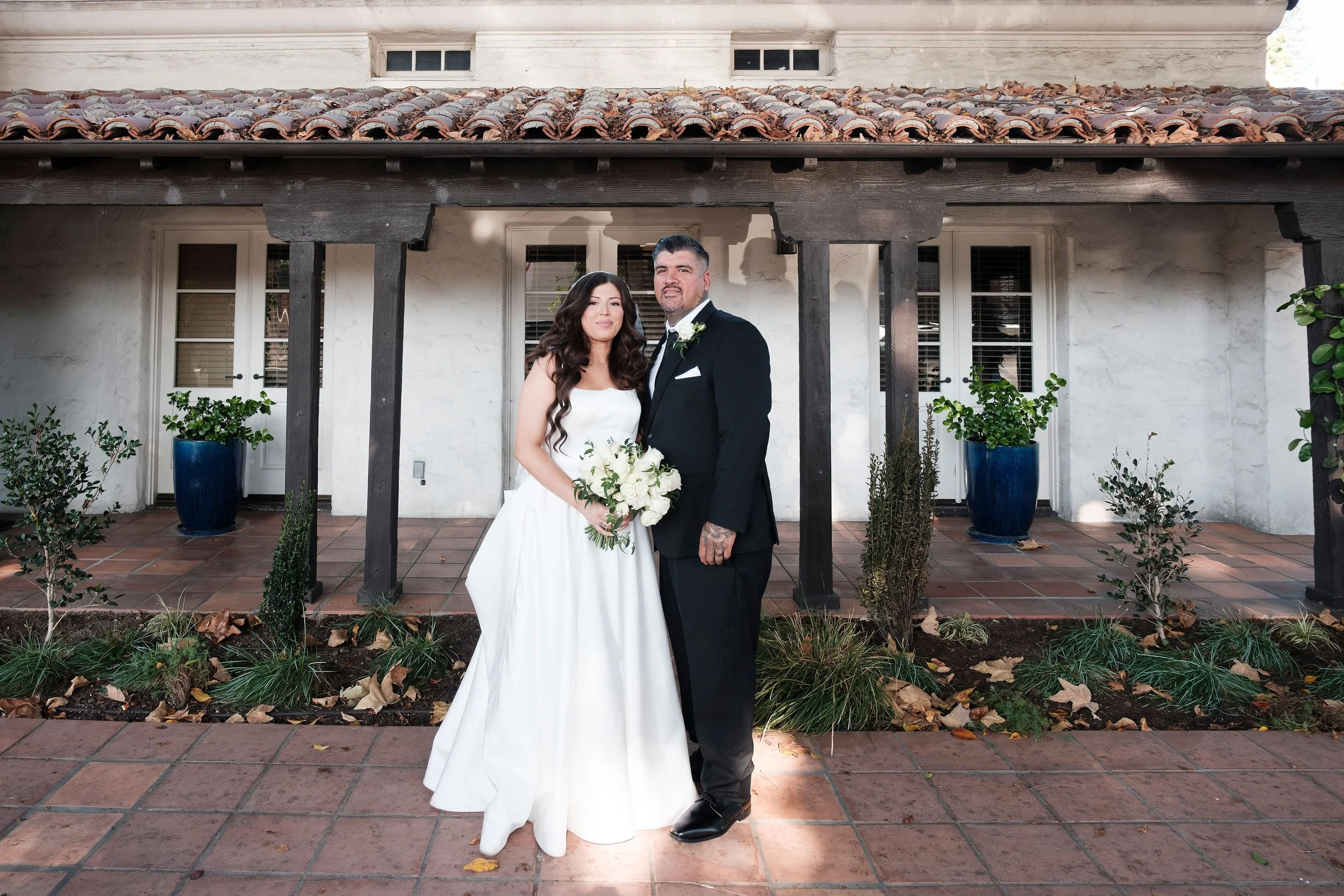 A newlywed couple standing outside in front of a white stucco house with wooden beams and tile roof. The bride is holding a bouquet of white roses, wearing a white wedding gown with long dark hair. The groom is dressed in a black tuxedo with a white shirt and boutonniere. They are posing together on a brick patio surrounded by potted plants and small bushes.