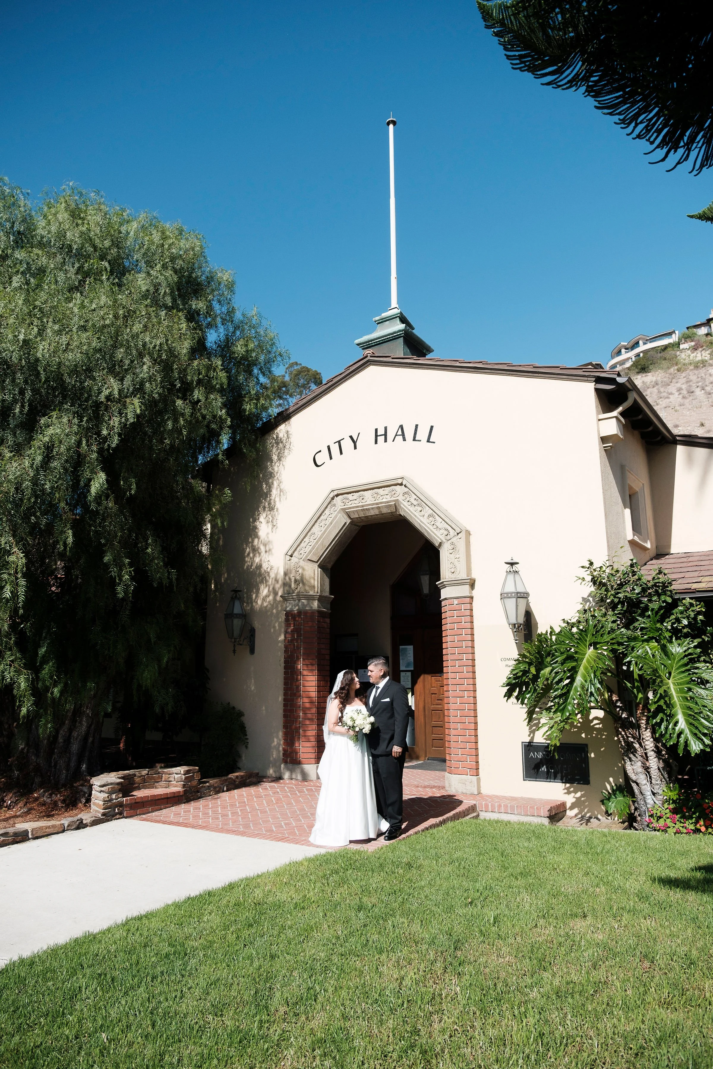 A bride and groom standing in front of City Hall, holding hands and gazing at each other on their wedding day. The building has an archway entrance, brick and stucco exterior, and is surrounded by green landscaping.