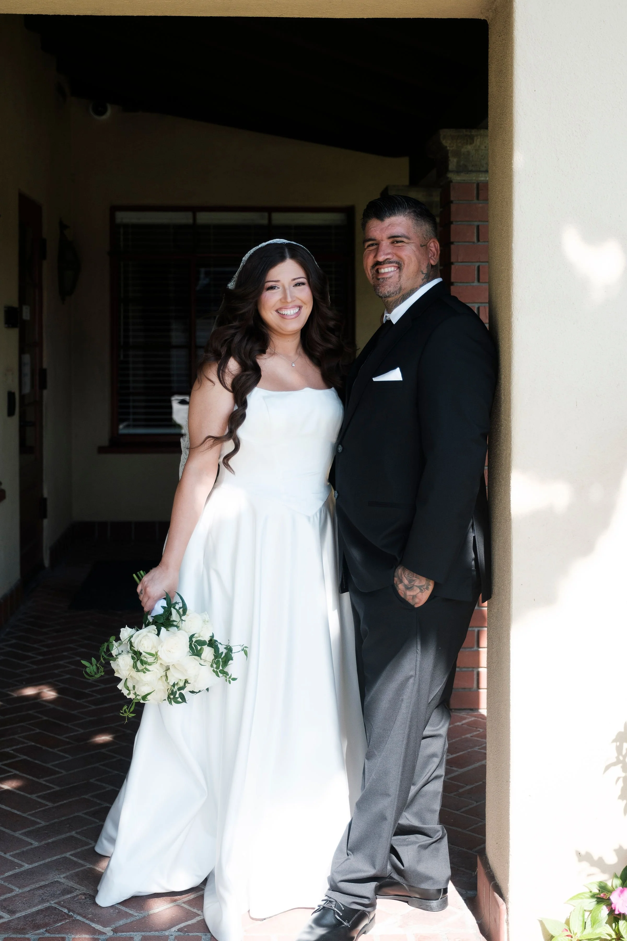 A bride and groom standing together outside a house, smiling. The bride is holding a bouquet of white roses and wearing a white wedding dress. The groom is dressed in a black suit with a white shirt and tie, leaning against a wall.