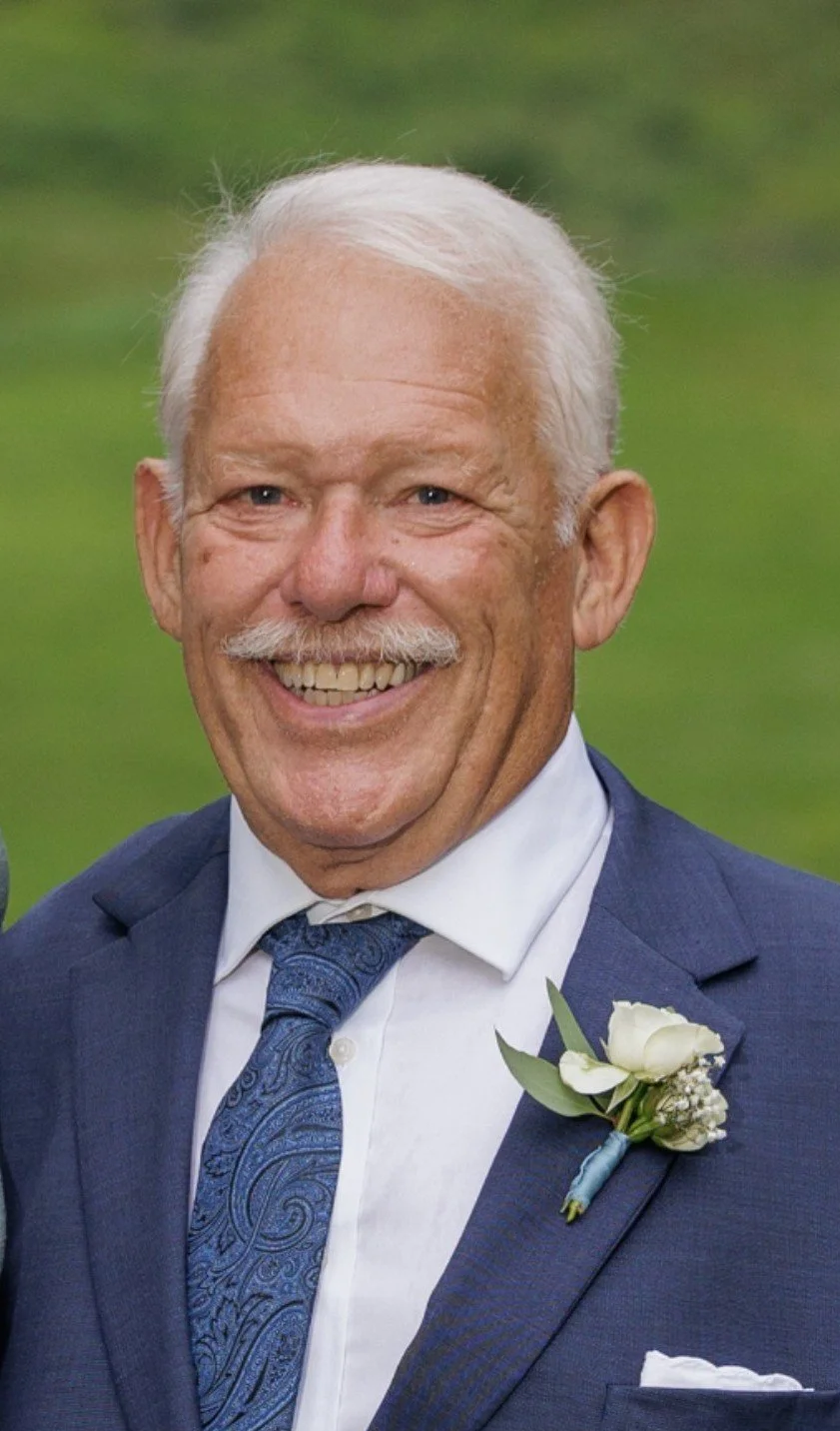 Portrait of an elderly man with white hair, a white mustache, wearing a blue suit, white shirt, and paisley tie, smiling outdoors with a blurred green background.