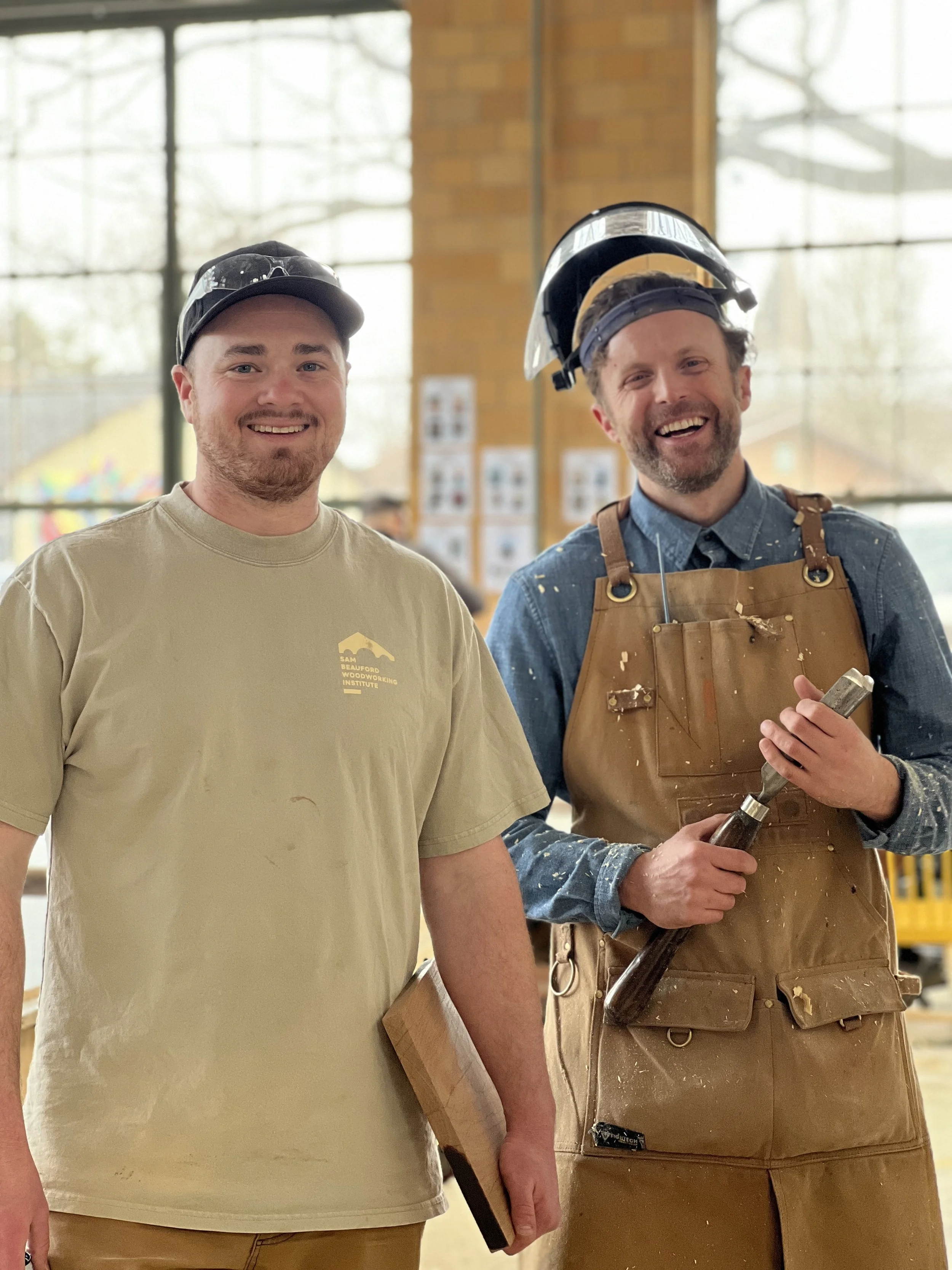 Two men standing inside a woodworking shop, one wearing a beige shirt with logo and the other in a denim shirt and apron, holding tools and wearing safety helmets, smiling.