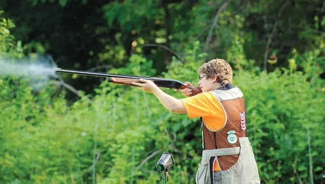 Young boy aiming a shotgun outdoors in a wooded area.