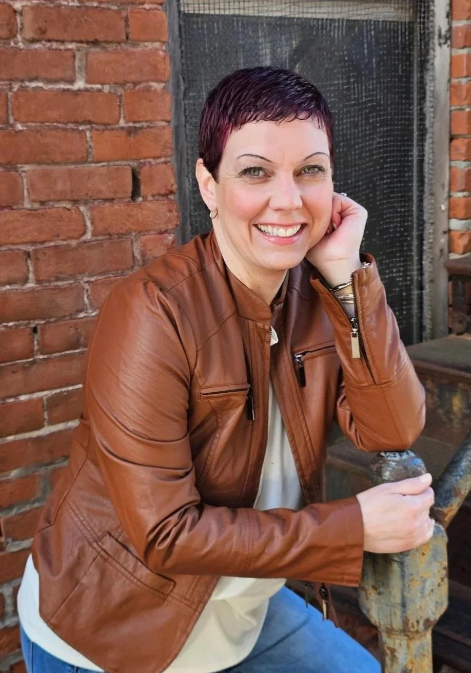 A smiling woman with short dark hair and earrings, wearing a brown leather jacket and white top, sitting outdoors against a brick wall.