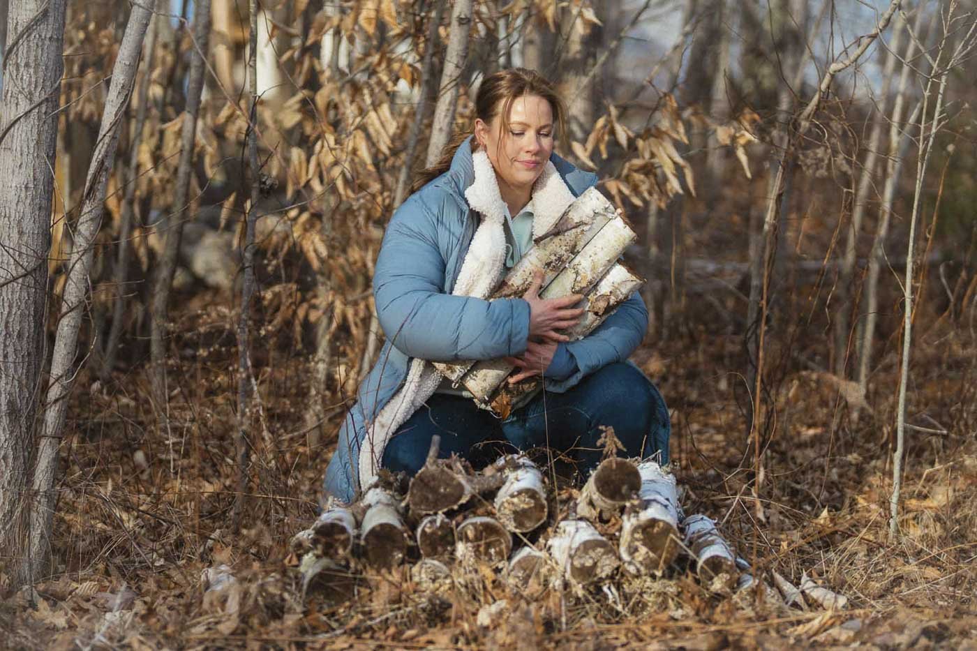 A woman kneeling in a forest during autumn, holding several logs of firewood. She is dressed warmly in a blue jacket and jeans, with fallen leaves on the ground.