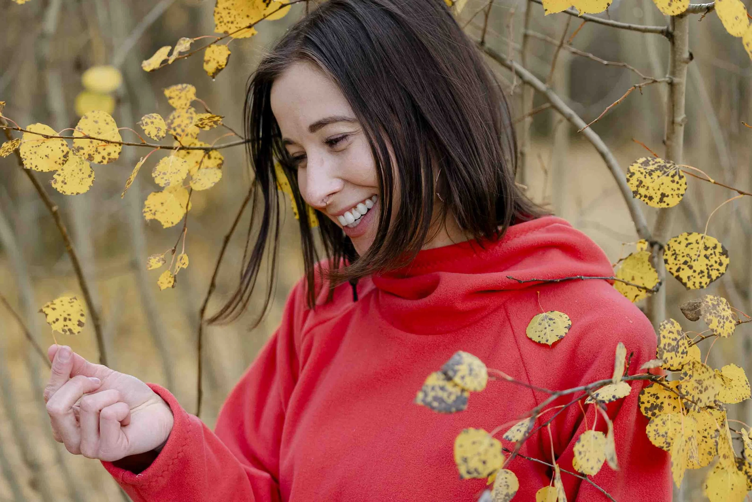 A woman in a red hoodie smiling and looking down while standing among yellow autumn leaves on a tree branch.