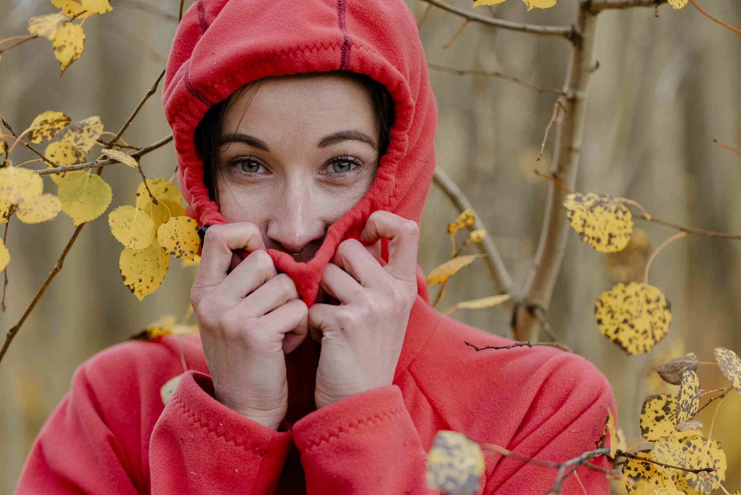 A woman wearing a red hoodie pulls the hood over her face, revealing her eyes and nose, amid autumn foliage in the background.