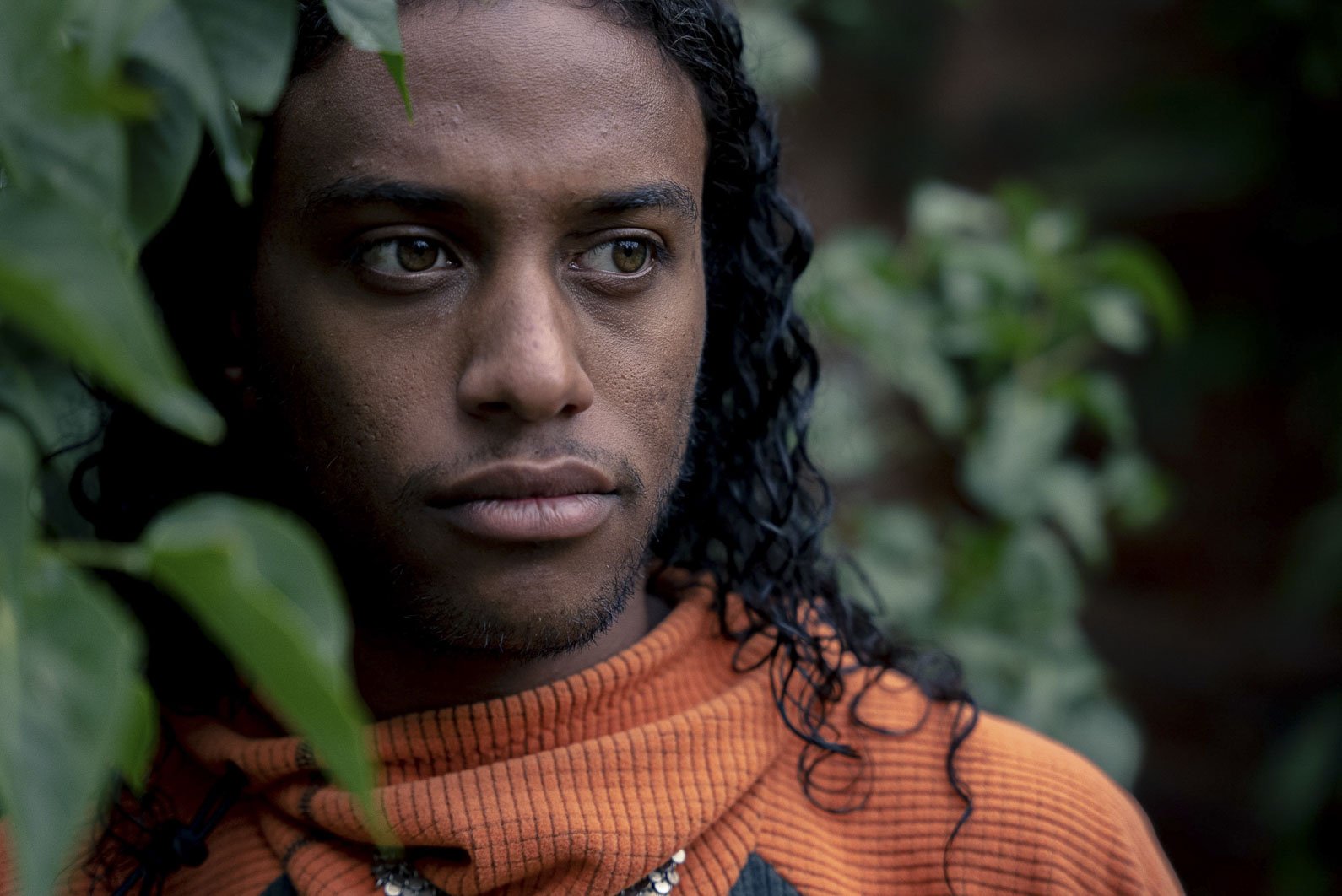 Close-up of a person with long, curly black hair and brown eyes, wearing a burnt orange sweater, surrounded by green foliage.