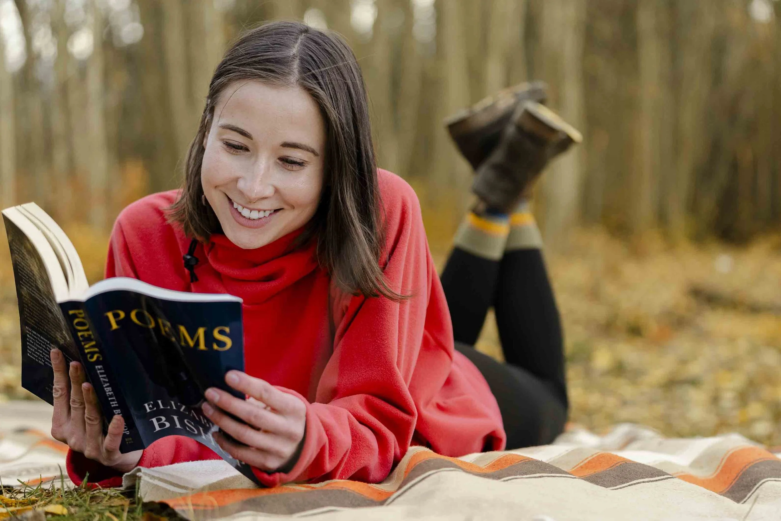 A woman lying on a blanket outdoors in a forest, reading a book and smiling