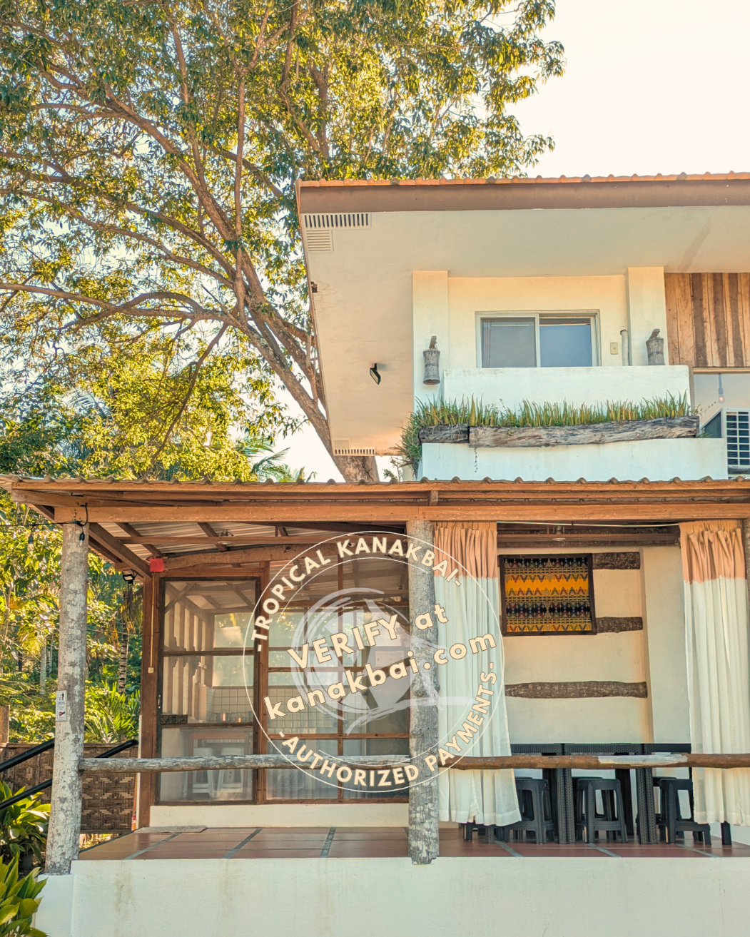 A tropical house in Mati City Dahican Beach with a balcony and sliding glass doors, surrounded by green trees, under a clear sky.