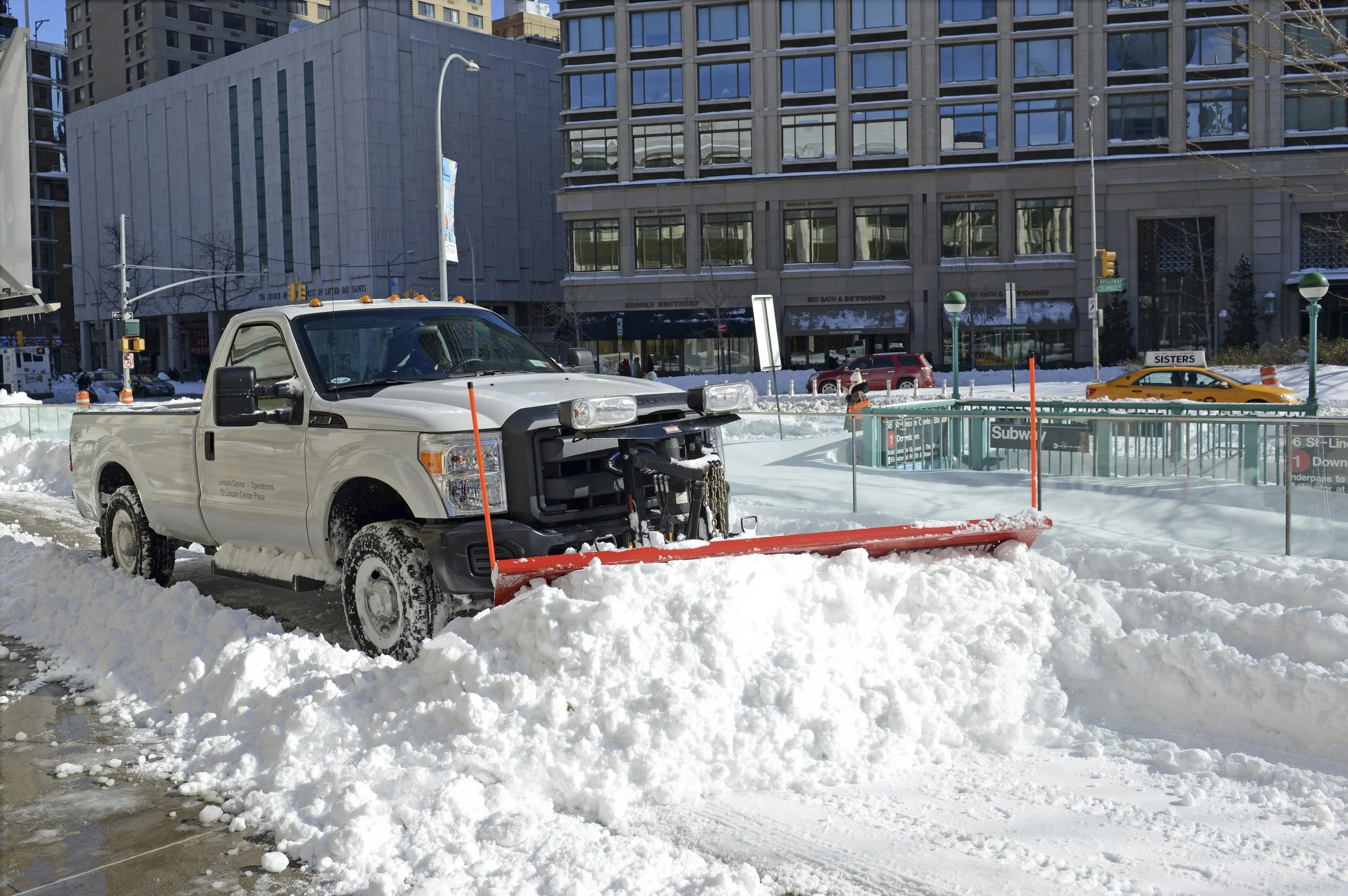 A snowplow clearing snow from a city street near a subway entrance in downtown, with tall buildings and parked cars in the background.