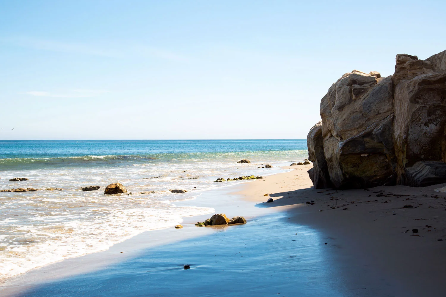A beach with sand, ocean waves, and large rocks on the right side under a clear blue sky. Photographed by Kayla Duffney Creative.