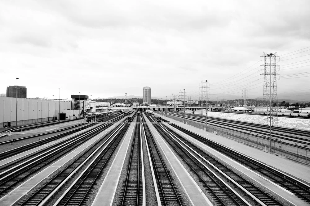 Black and white photo of multiple train tracks converging towards the horizon with a city skyline in the background under cloudy sky. Photographed by Kayla Duffney Creative.
