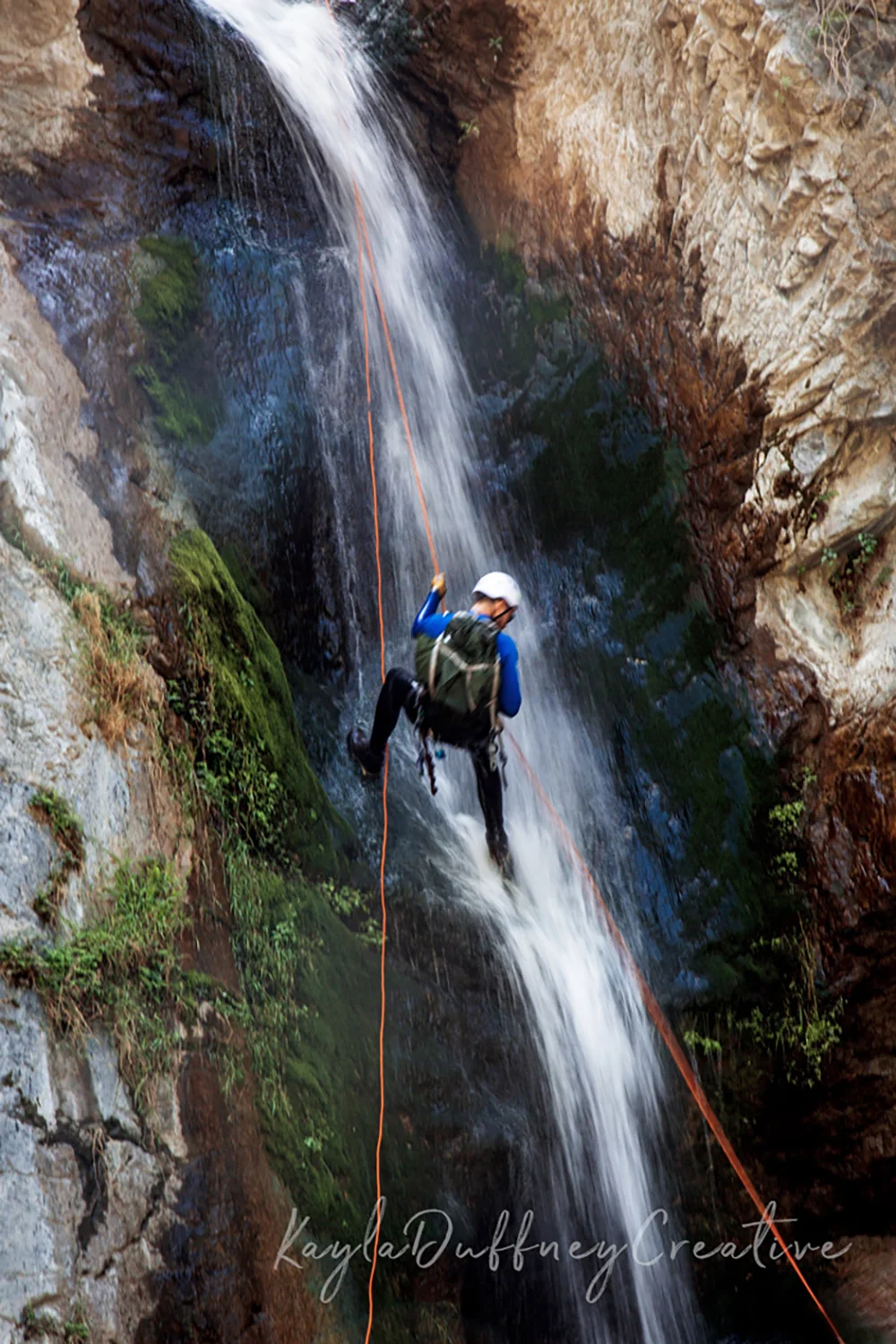 A person in climbing gear rappelling down a waterfall in a narrow canyon with moss and rocks. Photographed by Kayla Duffney Creative.