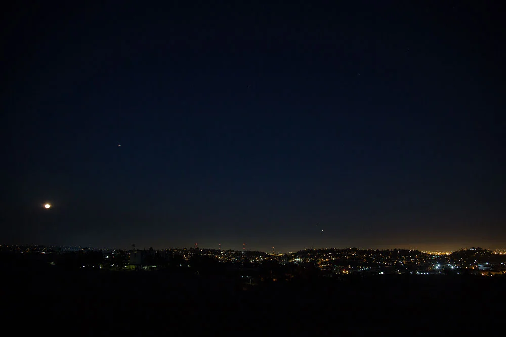 Nighttime cityscape with a bright moon and stars in the dark sky over a city with illuminated buildings. Photographed by Kayla Duffney Creative.