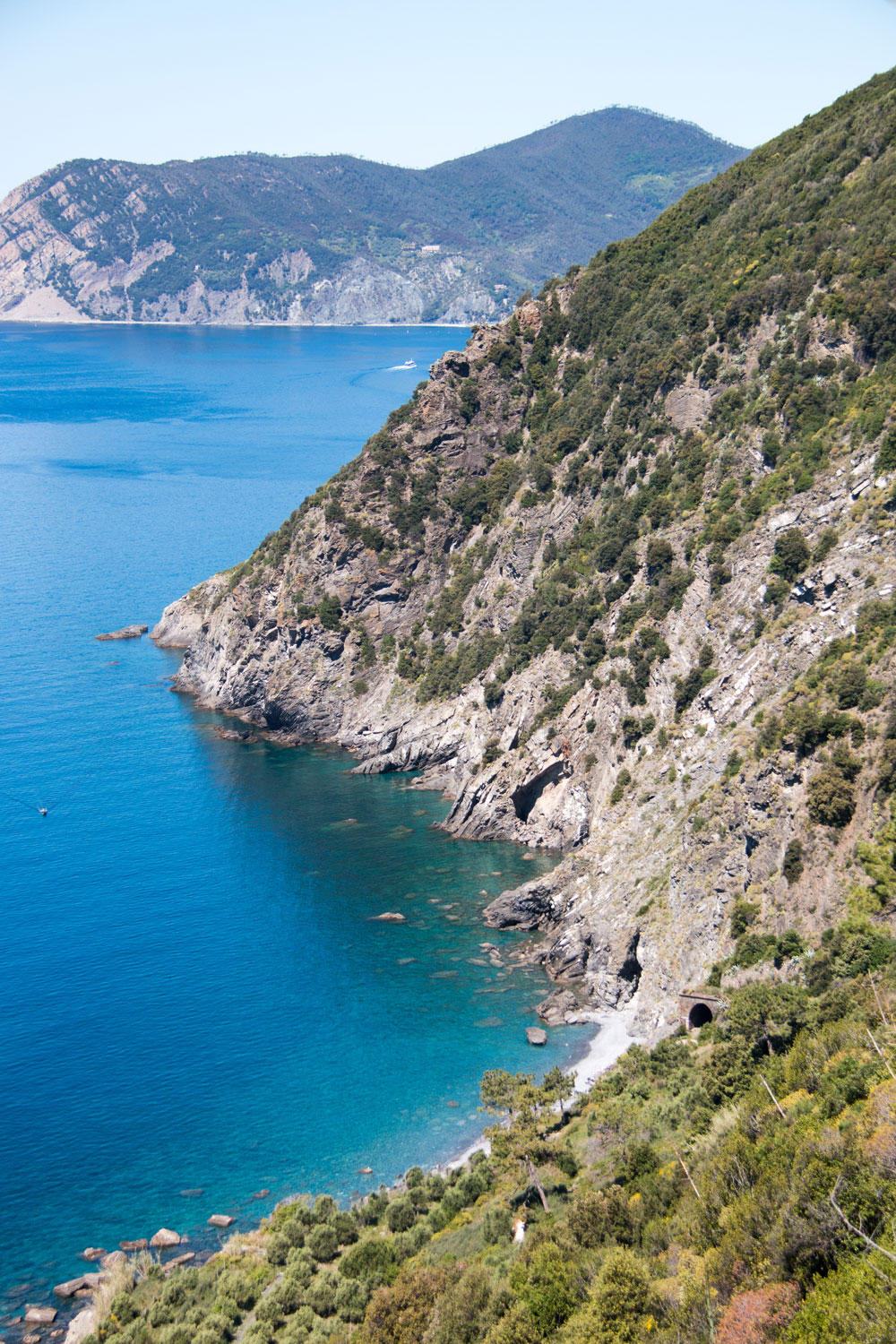 Coastal view of steep, rocky cliffs covered in greenery beside a calm, blue sea with a small beach and a tunnel in the hillside. Photographed by Kayla Duffney Creative.