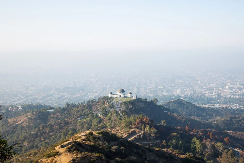 The Griffith Observatory atop a hill overlooking Los Angeles with the cityscape in the background under a hazy sky. Photographed by Kayla Duffney Creative.