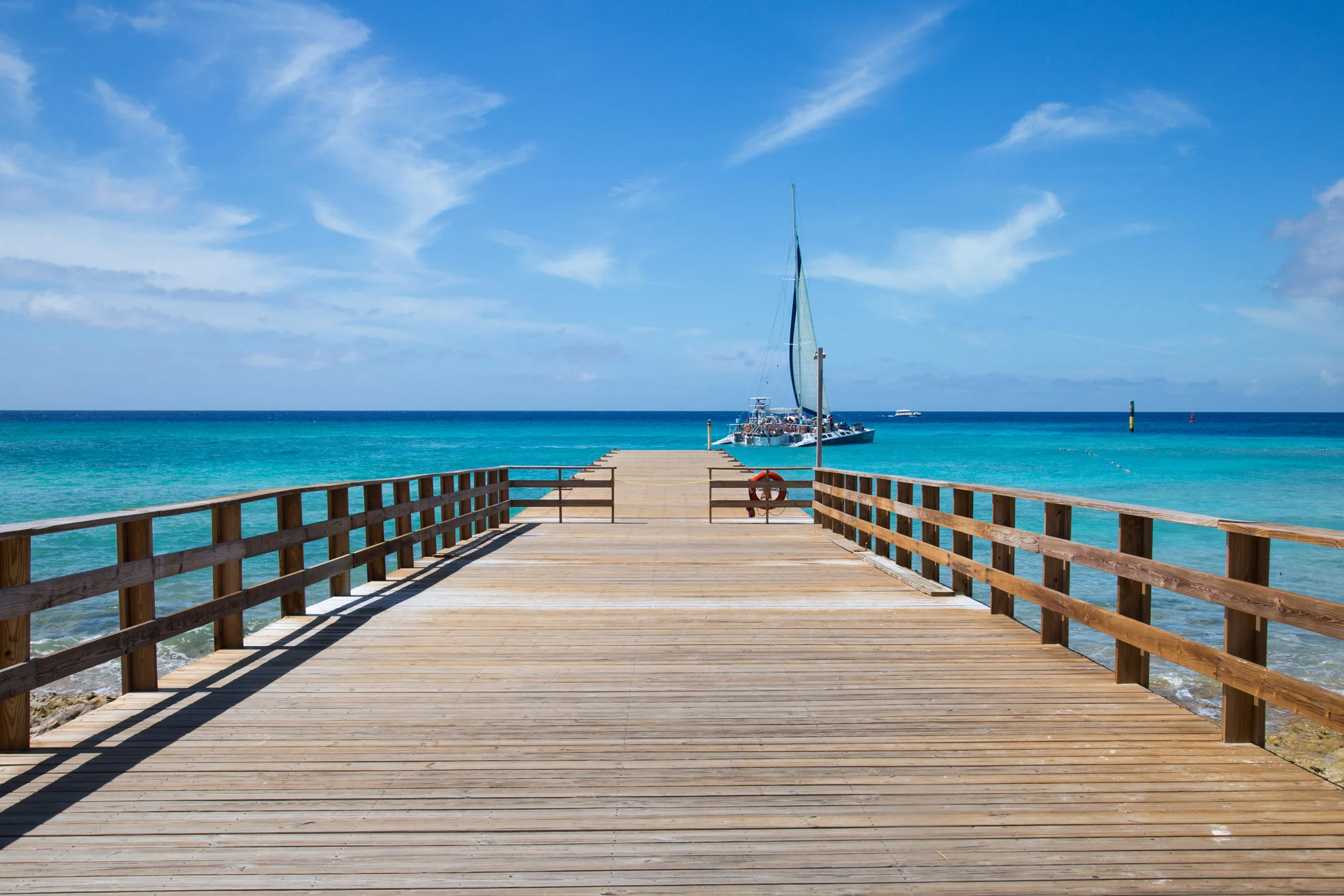 A wooden dock extends into the turquoise ocean under a blue sky with wispy clouds. A sailboat with sails down is anchored near the dock, and a boat is visible on the horizon. Photographed by Kayla Duffney Creative.