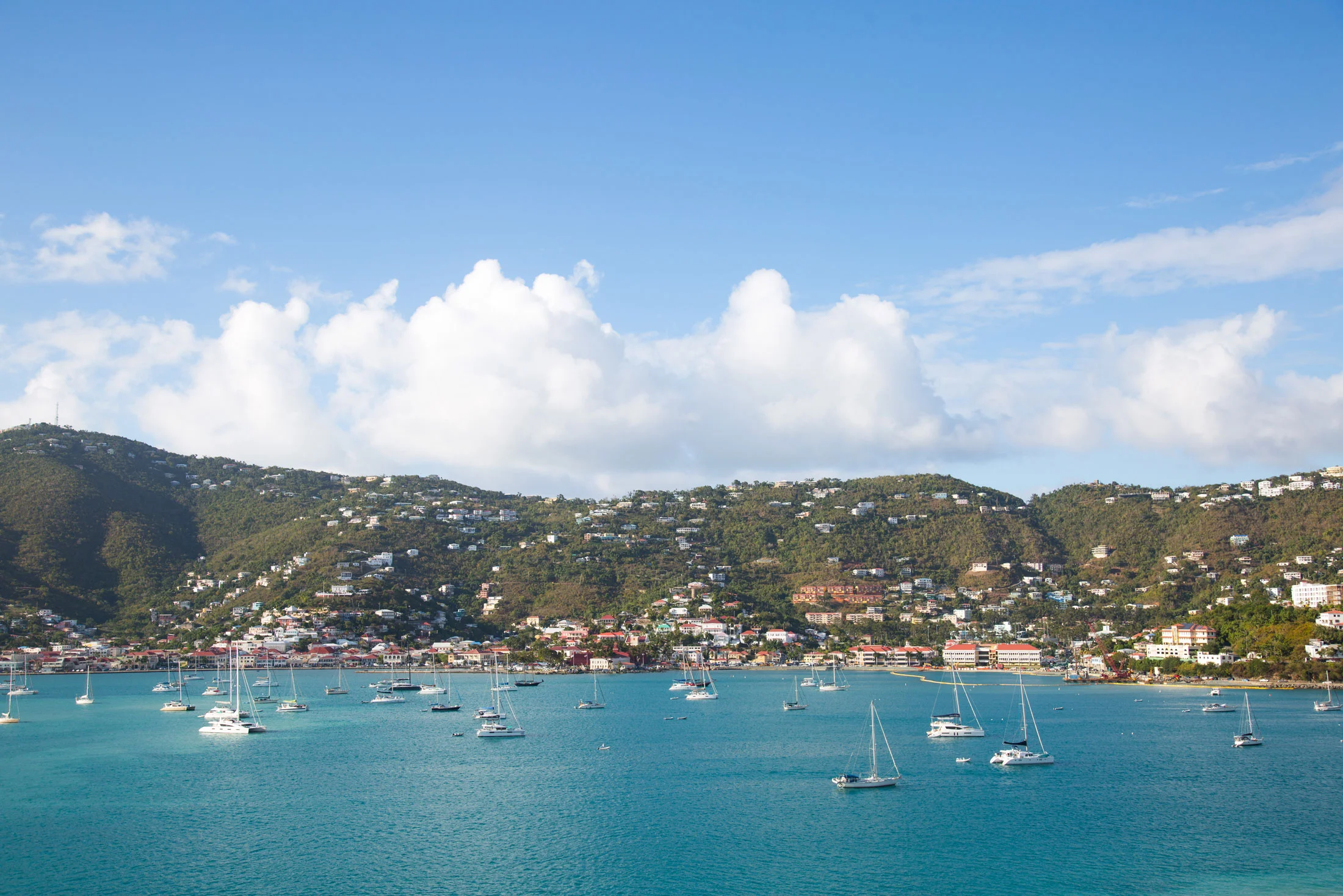 View of a harbor with sailboats in the turquoise water, hillside with houses, and a blue sky with clouds. Photographed by Kayla Duffney Creative.