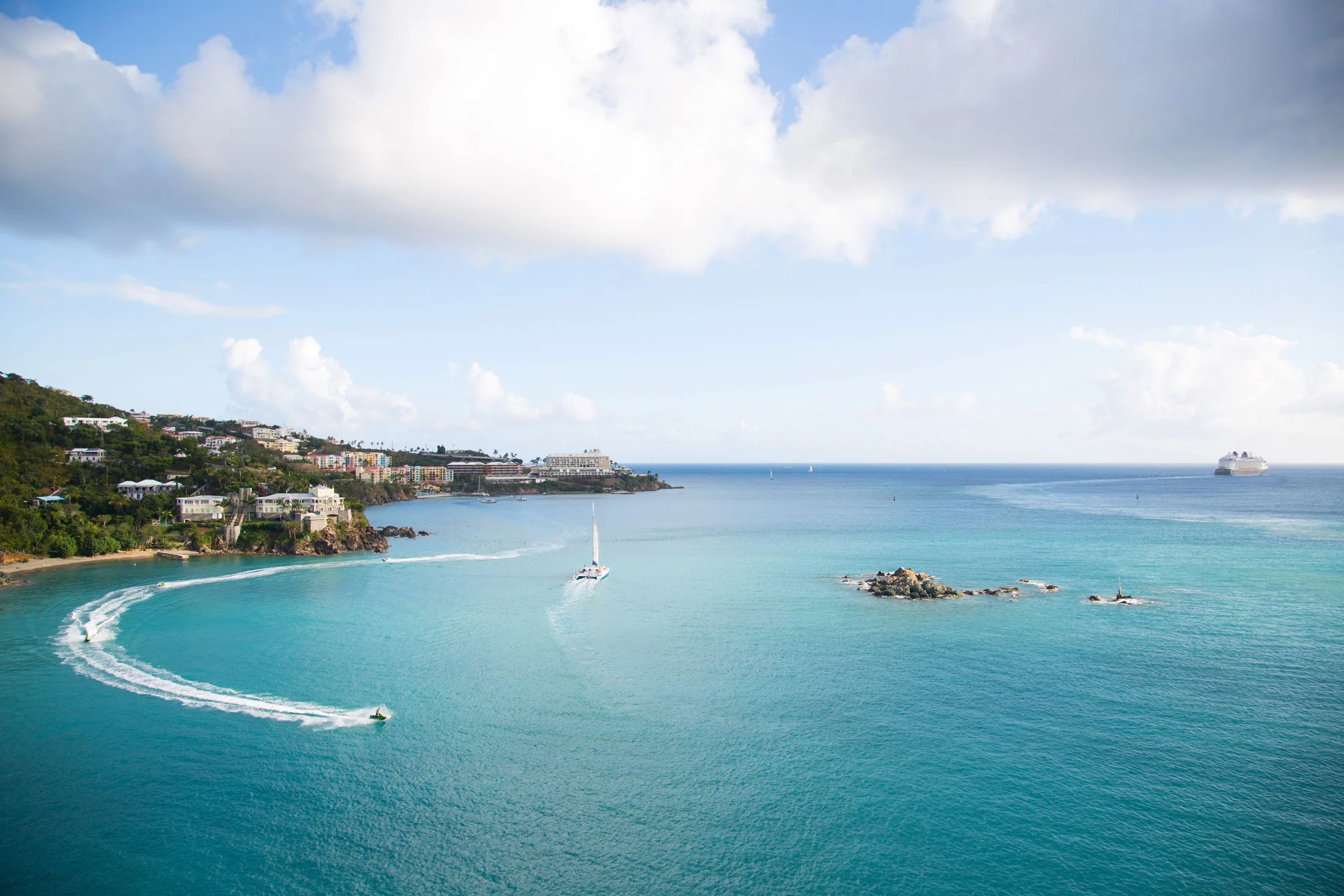 Aerial view of a coastal area with boats on turquoise waters, hillside with houses, and a cruise ship in the distance under a partly cloudy sky. Photographed by Kayla Duffney Creative.