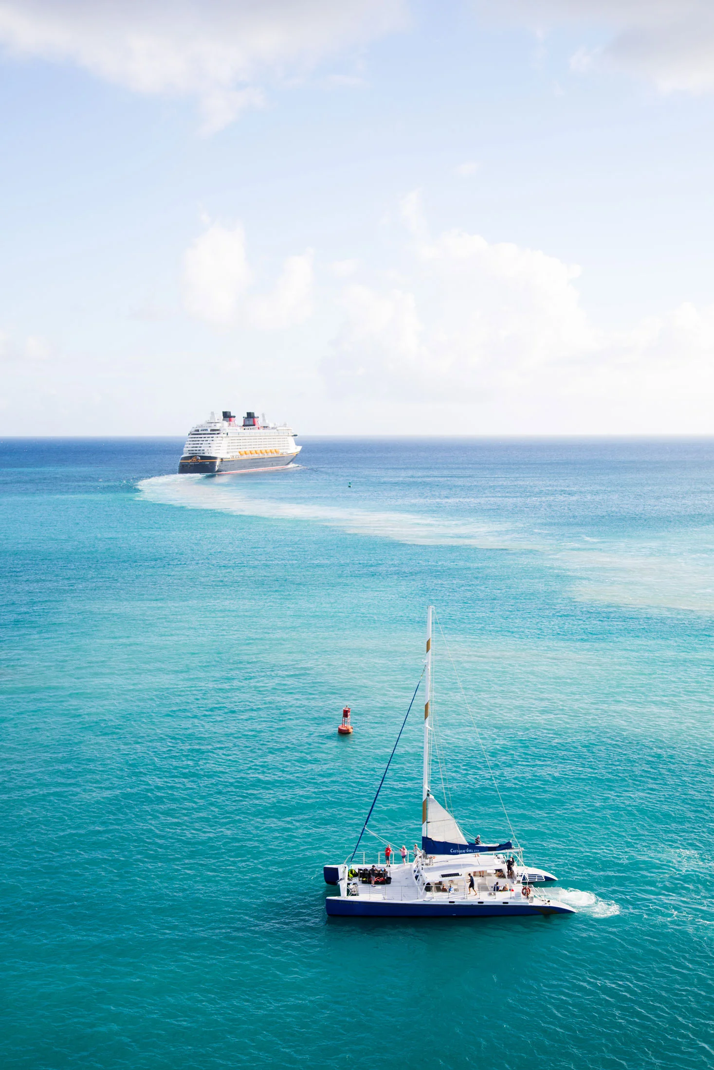 A white catamaran sailboat with a blue sail in clear turquoise waters, with a cruise ship in the distance under a partly cloudy sky. Photographed by Kayla Duffney Creative.
