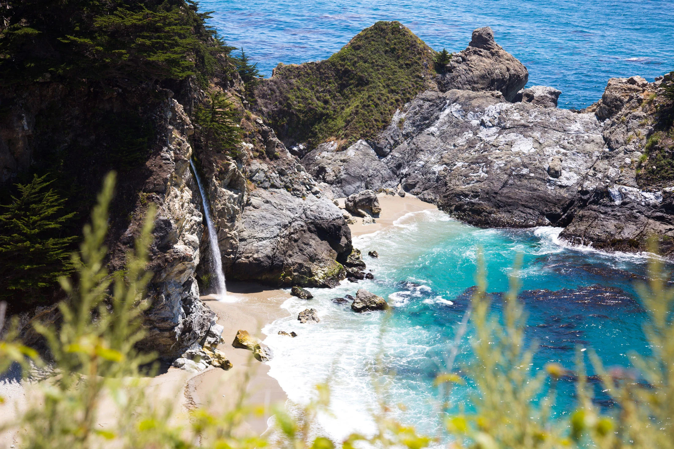 A coastal scene with rocky cliffs covered in green vegetation, a small sandy beach, a waterfall, and blue ocean waters with waves. Photographed by Kayla Duffney Creative.