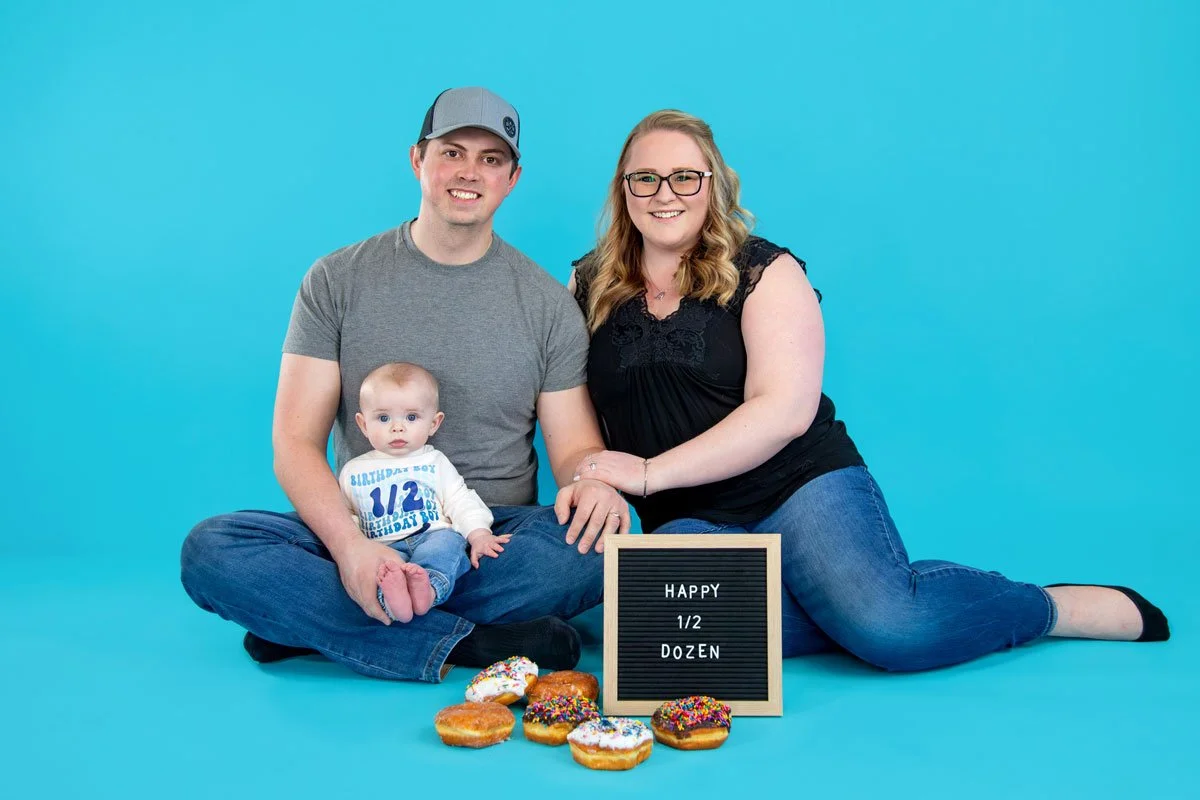 A family of three sitting on the floor against a blue background, celebrating a 6-month-old's birthday with a sign that reads "HAPPY 1/2 DOZEN" and a spread of donuts topped with colorful sprinkles.