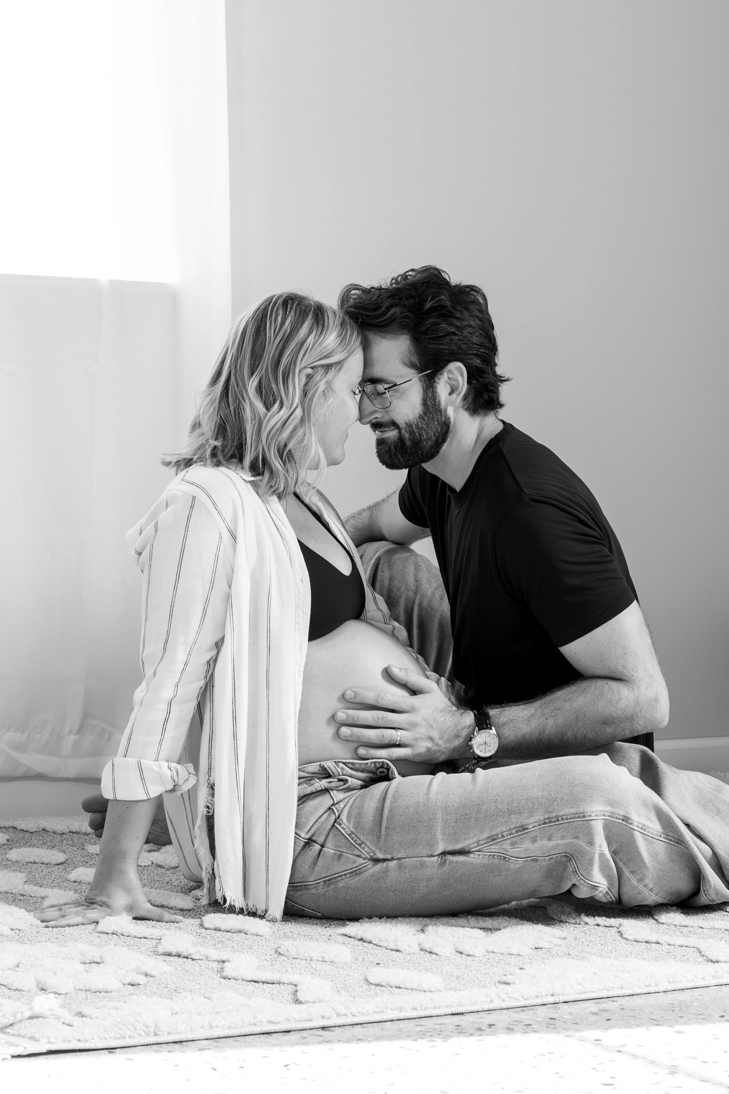 A pregnant woman sitting on a blanket on the floor with her partner kneeling in front of her, touching her belly. They are touching foreheads and smiling with their eyes closed. The woman is wearing a striped shirt and the man is wearing glasses and 