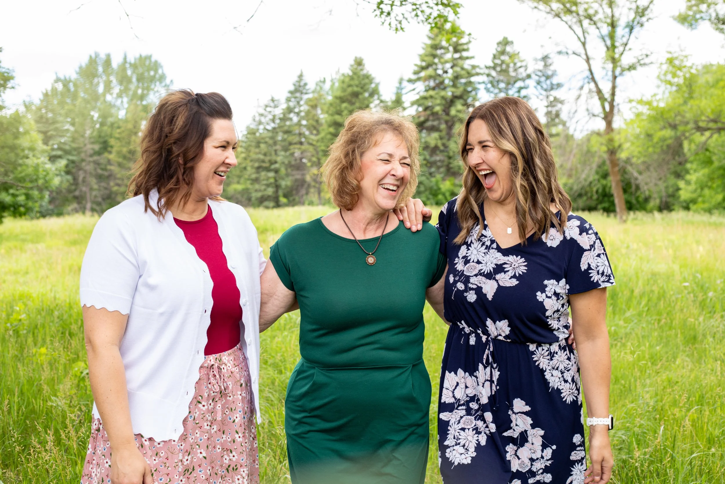 Three women smiling and laughing outdoors in a green, wooded area with tall grass.