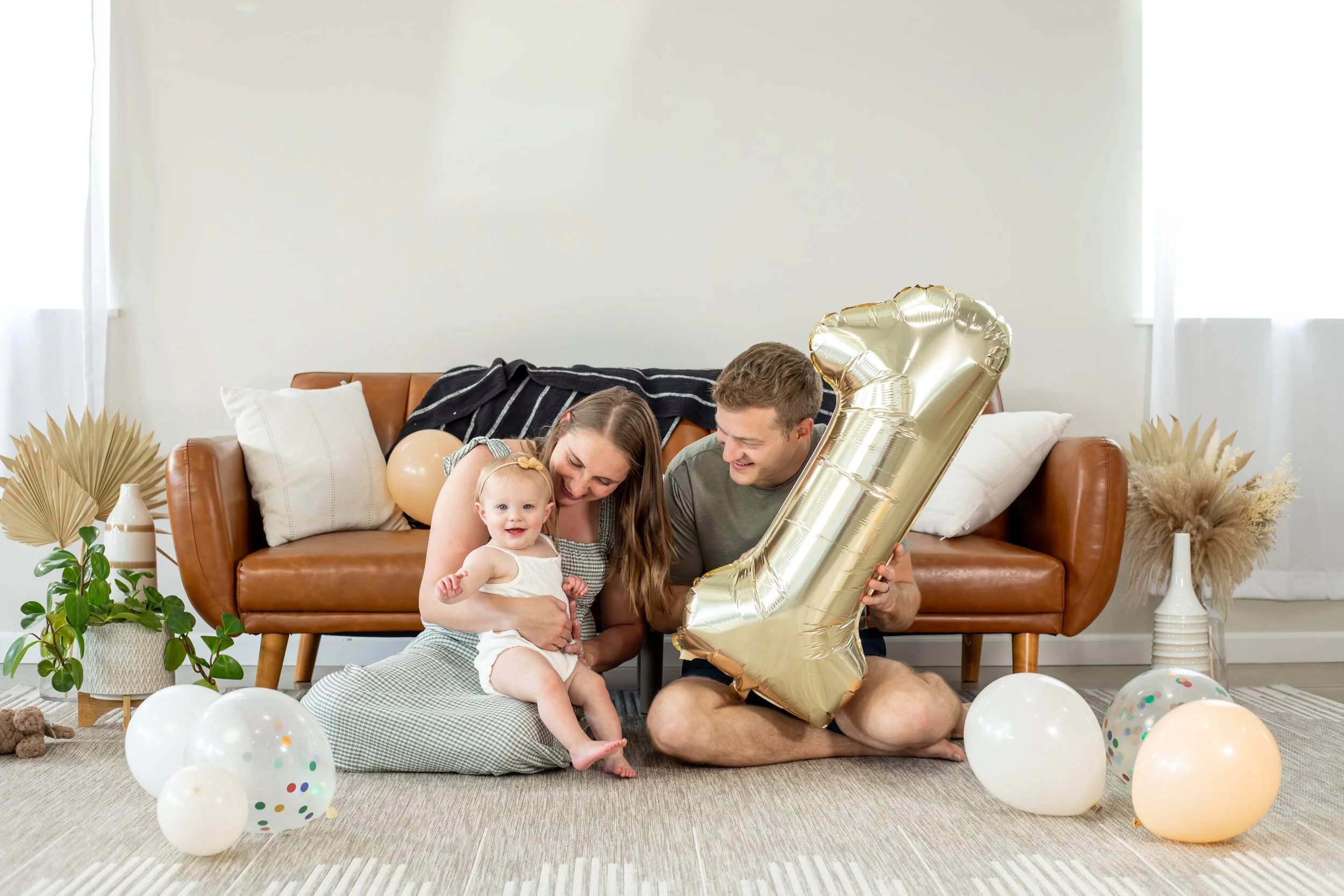 A family celebration scene with a smiling family of four sitting on the floor amidst colorful balloons, with the father holding a large gold number 1 balloon, in a cozy living room with a tan leather couch, white pillows, and decorative plants.