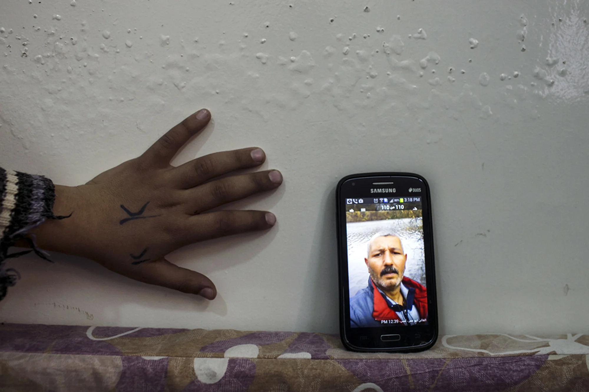 Jordan, 2014, Muna, a Syrian "urban refugee" and female head of house shares photographs of her husband from Germany. 'I swear each photo of him standing in the sun, or by an old church...you see devastation and guilt on his face. He wants to be with us so badly. We survive off of this photos and the lullabies and love messages he sends us on whatsap.' Her daughter's hand reaches for phone. 