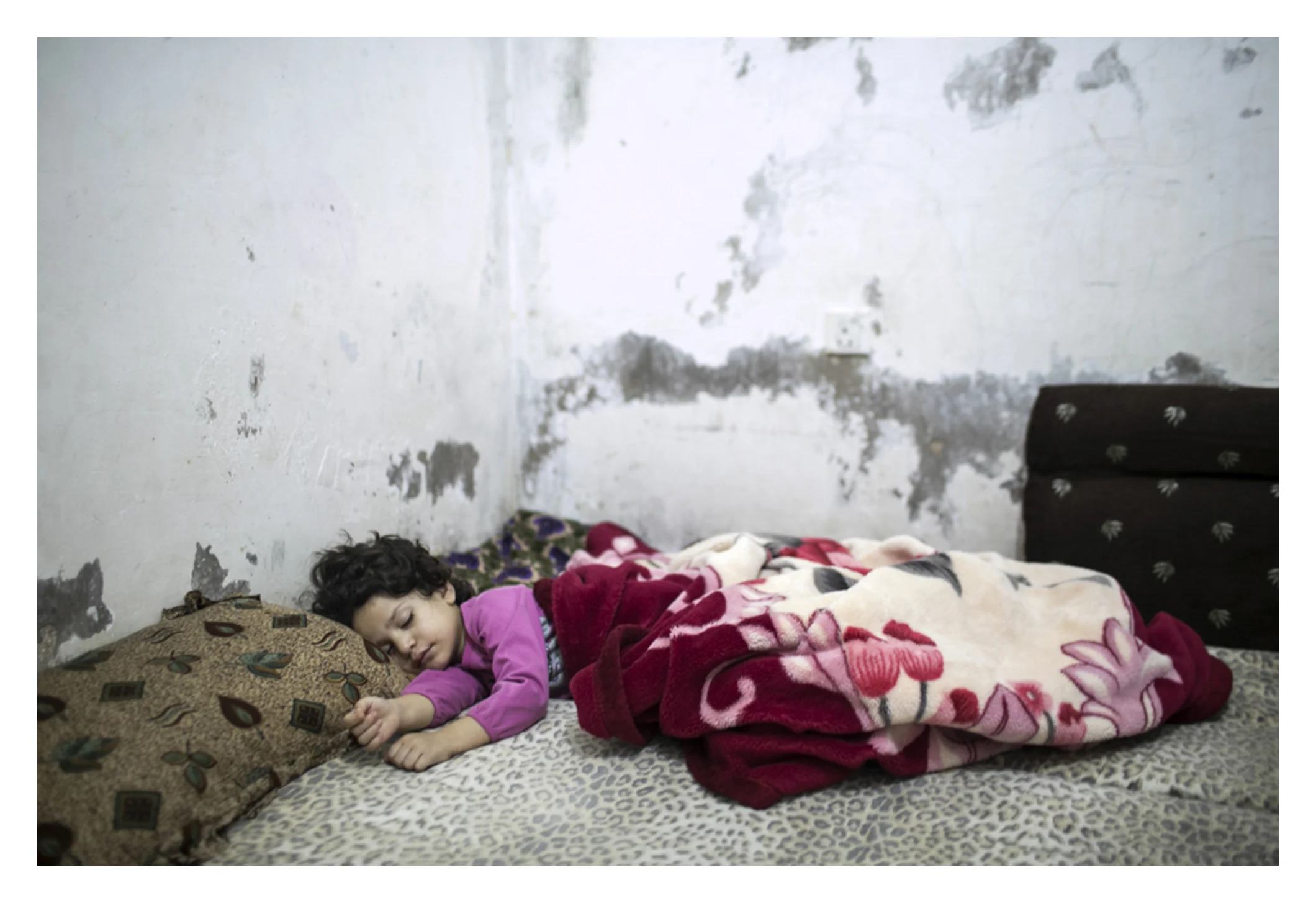 Five year old urban refugee reluctantly awakes to start her day.
An urban refugee family (refugee from Syria) are struggling to survive. Their father, Maher, 43, was paralyzed after he received an injection while he was in prison. Both the mother, Farah, has thalassemia, and their youngest daughter. UNHCR cleared them for cash assistance, which will be a lifesaver for the family. They pay 150 Jordanian dinars a month for a small, cold, and leaking apartment. Their only form of income is from the mother (she earns five Jordanian dinars a day as a maid) and from their 14-year-old son, Yazan. Yazan is 14 with a heart birth defect. He works at a bakery, a distance of half an hour walk, working from 7 till 10 at night for 7 JDs a day. ‘I can’t bear my mother working as a maid,’ Yazan says. He cannot find time to work in school, and returns home so exhausted his family watch him sleep and cry, they say. Their 12-year-old daughter Lamis helps with the house chores when her mother goes to work. She goes to school at 12:30, and comes back at 4:30. Through her work cleaning at the institution, Farah has secured a position at a private day care for her youngest daughter. Farah walks every day in the street looking for a job, asking if people need help in the house for cleaning.