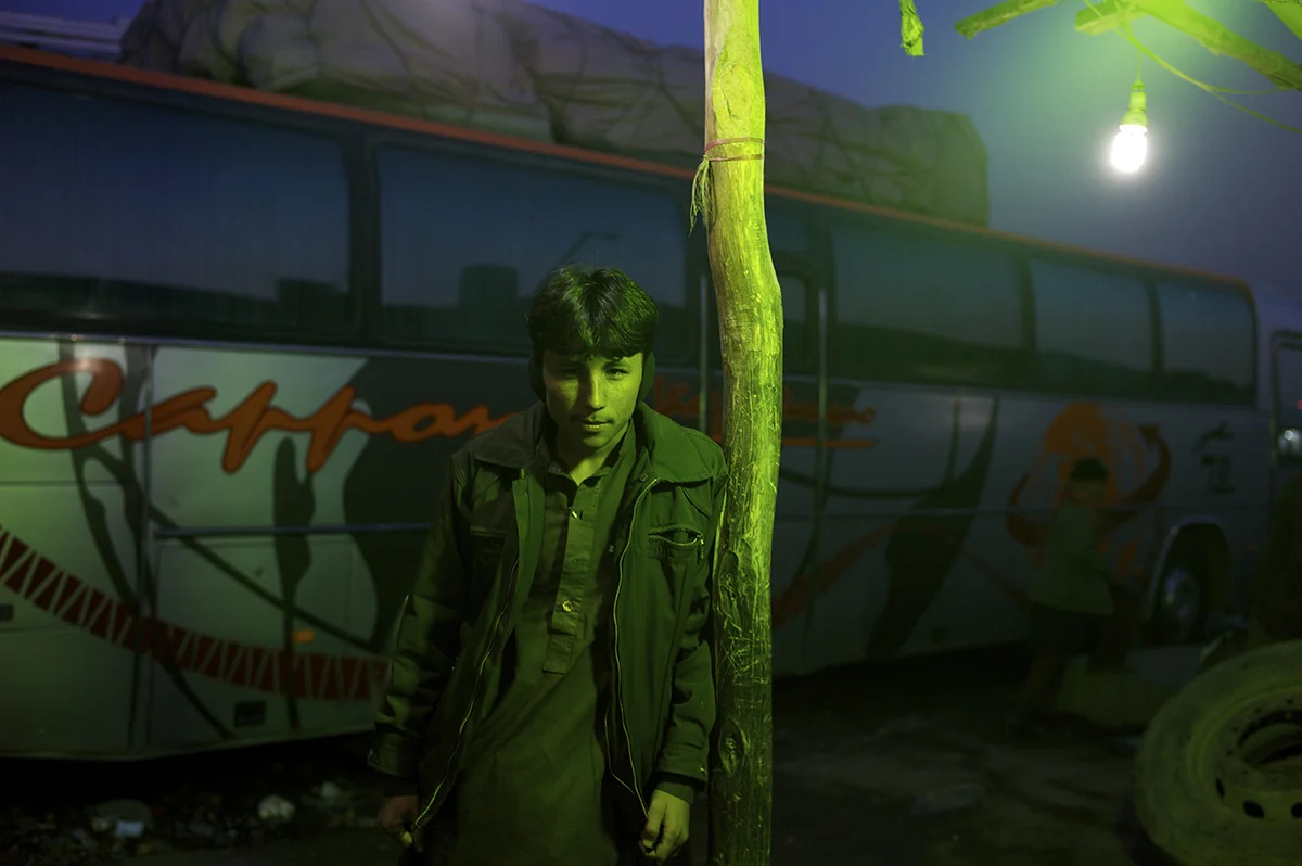 Afghanistan, Kabul, December 2011, Under the green fluorescent lights of a shelter, a teenage traveller in ear muffs waits outside a mosafer khana in the grounds of the Company area bus station.