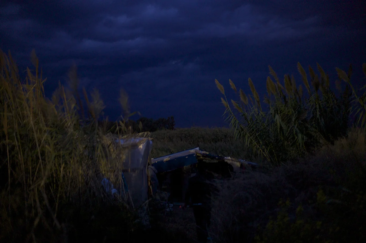 Greece, Patras, October 2011, Strong winds blow through the pampas grasses surrounding a crudely built shelter just illuminated by the glow from the nearby Silk Road petrol station at night, at a spot known locally to Afghans as Pump Benzene Camp.