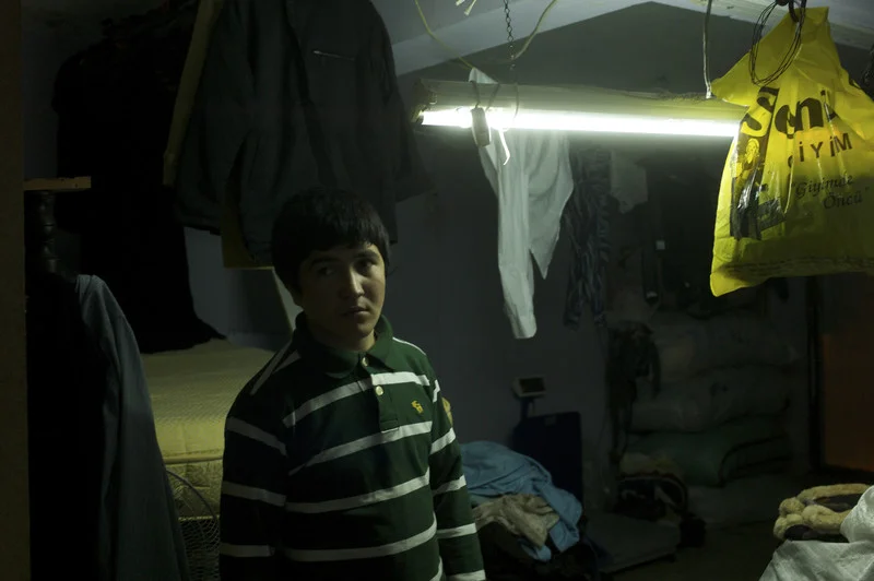 Turkey, Istanbul, January 2012, Next to a mattress balanced over the basement stairs, fourteen year old Abdul Hashim stands amid the cluttered storeroom in the kargah where he has spent the past months cutting cloth.
