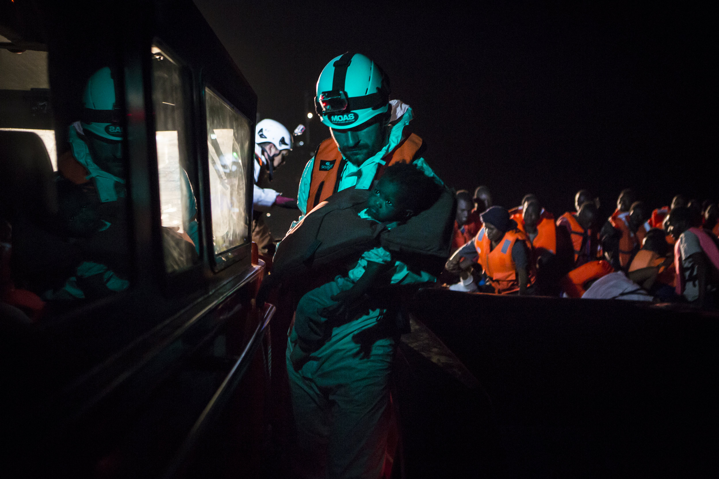  A MOAS rescue team member carries a baby from the rubber boat in the background to the safety of the rescue vessel. � Mathieu Willcocks/MOAS.eu 2016, all rights reserved. 