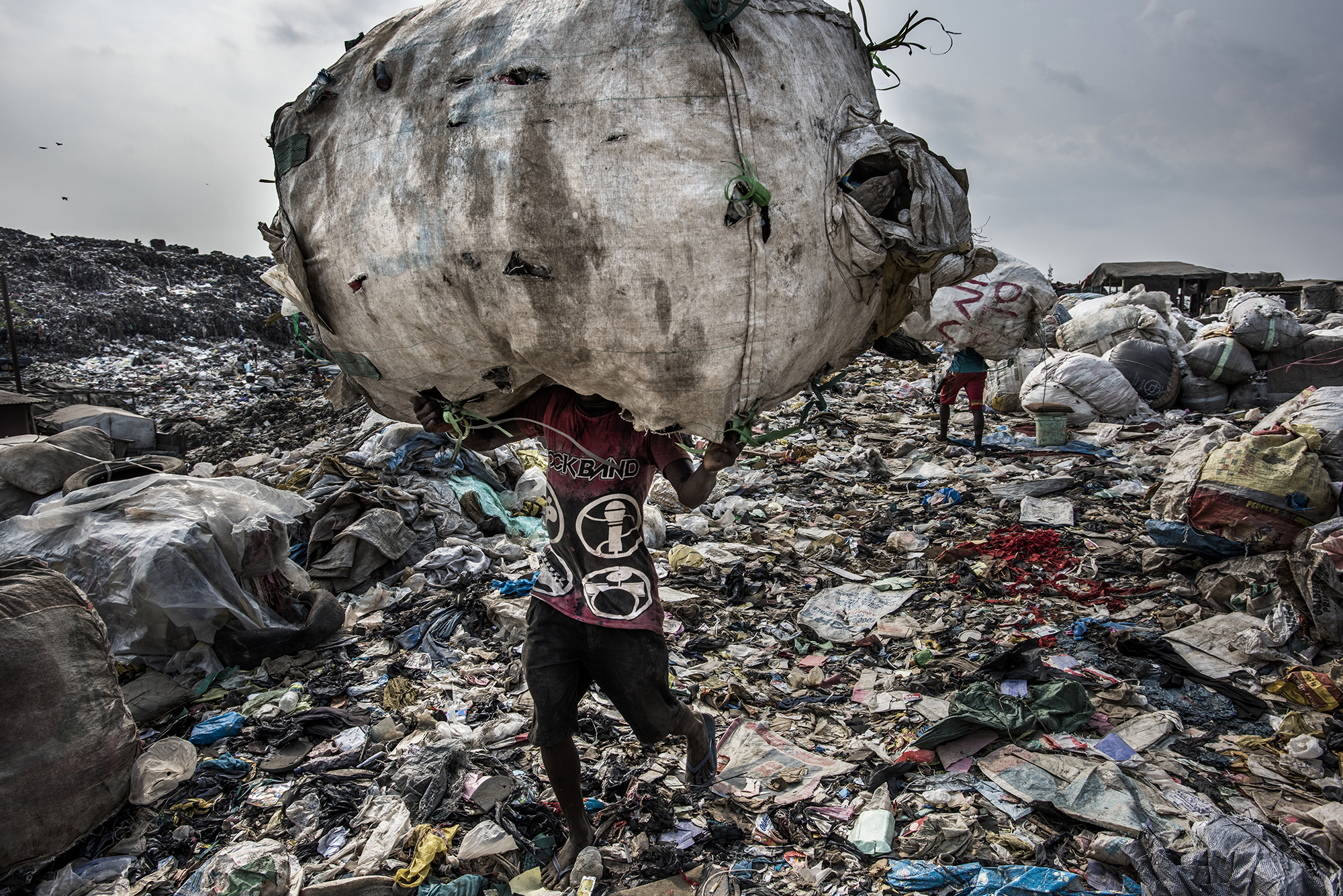 Nigeria, Lagos, 27 January 2017
A man carries a huge back of pet bottles collected for recycling at the Olusosun landfill.
The Olusosun landfill in Lagos receives between 3-5000 tons per day and is about 45 ha in size. About 5000 scavengers work here and often also live. They collect anything that is recyable like plastics, textiles, electronics, paper etc. The problem is that the landfill is full and the city wants to close it down. The question is where it will go, there are no incinerators and the infrastructure to formally recycle is lacking. There is one other landfill, but it needs to close as well.
Remarkable is that the landfills in Lagos smell less compared to other landfills in the world: Nigerians throw away less food, because they either finish their plate or feed it to the animals.
Kadir van Lohuizen / NOOR