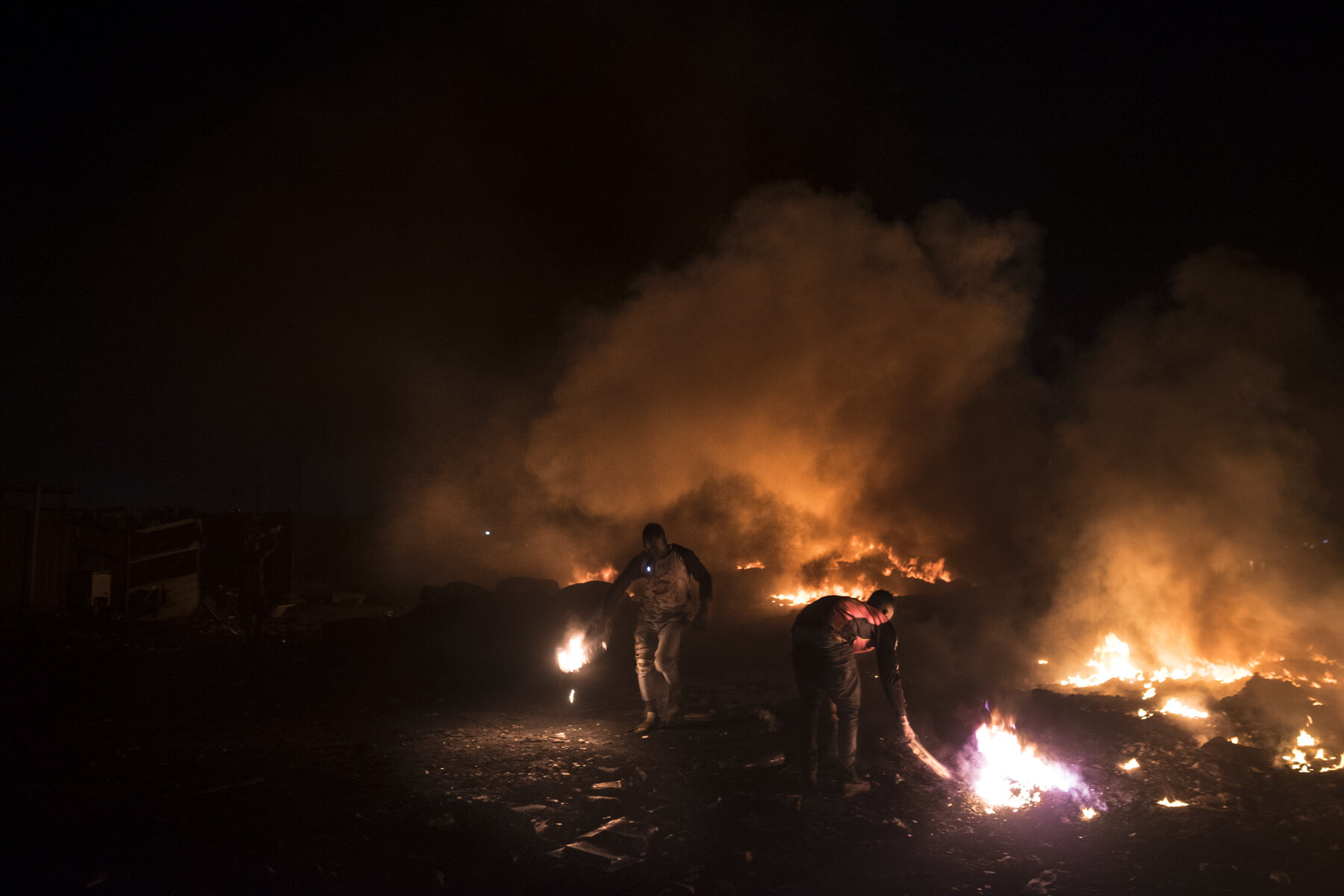  Ghana, Accra, November 2018.  Two workers burning cables, tires and parts of electrical appliances at night in the Kilimanjaro burning area of the Agbogbloshie scrap yard. Big fires are set at night to burn and extract as much raw material as possib